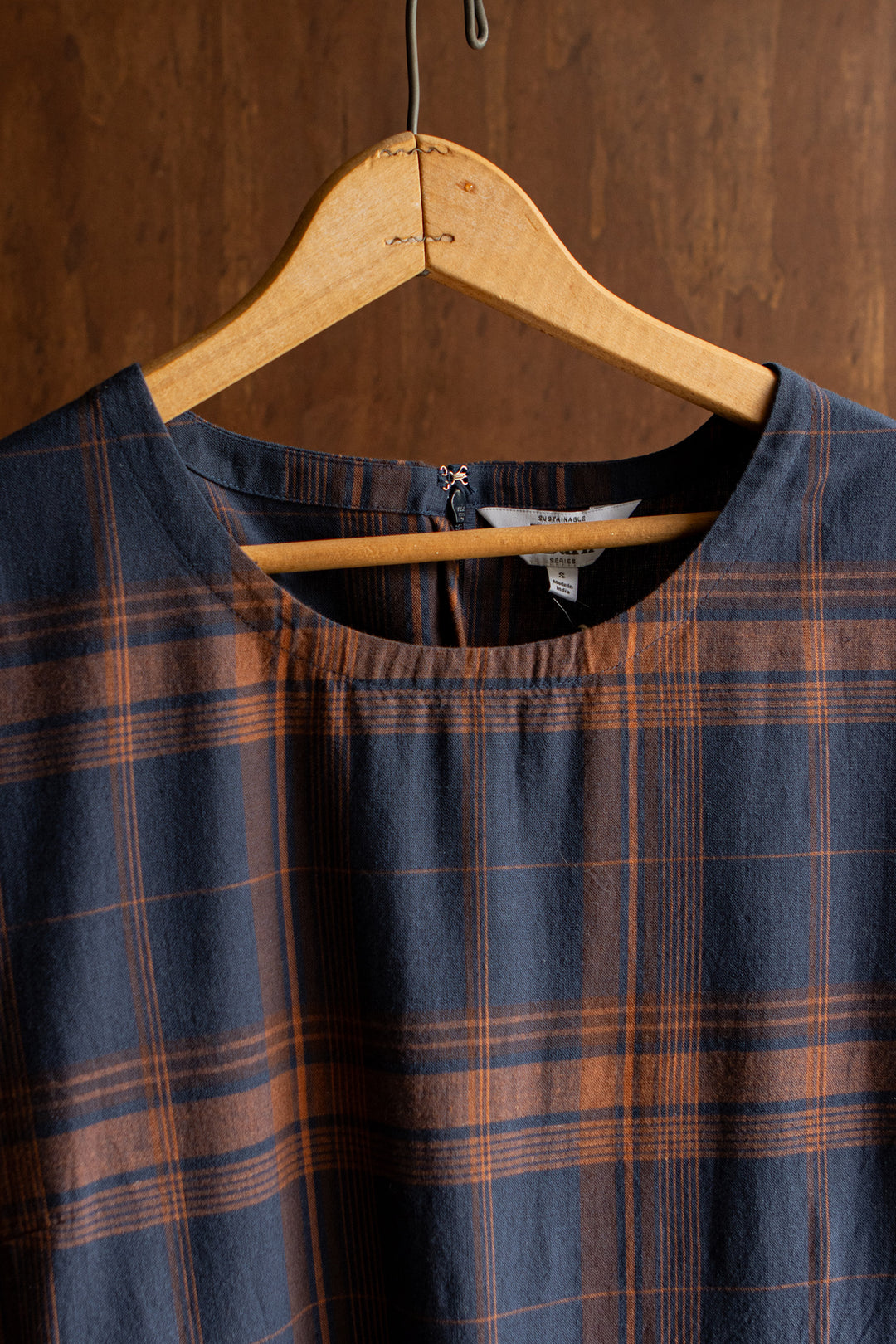 A close-up of the Bridge & Burn Perry Dress in Cinder Plaid—an organic cotton dress with a gathered skirt—hanging on a wooden hanger against a wooden background.
