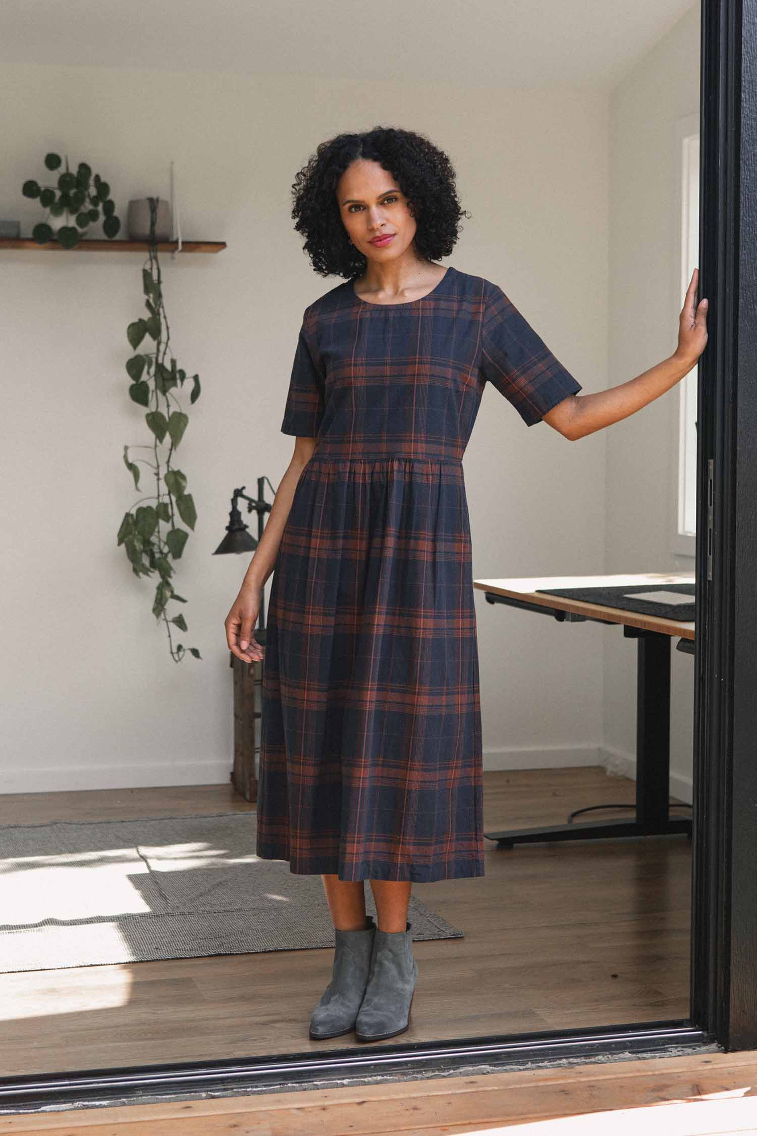 A woman wearing the Bridge & Burn Perry Dress in Cinder Plaid, paired with gray boots, stands in a bright room with wooden floors, a table, and a hanging plant visible in the background.