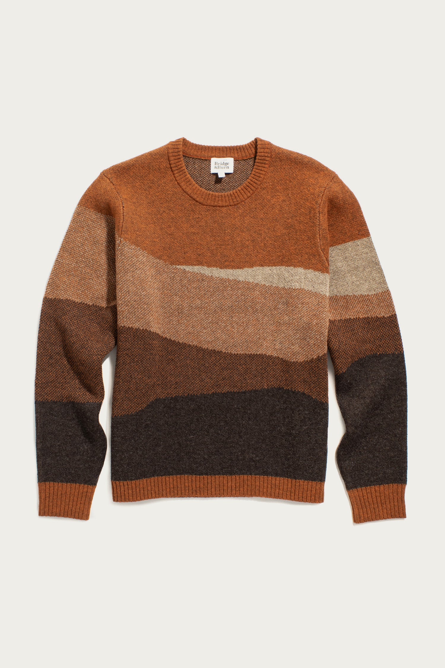 The Petra Sweater / Sandstone by Bridge & Burn is a crewneck with abstract, earthy stripes in brown, tan, rust, and black. Made from RWS-certified wool, it has long sleeves plus ribbed collar, cuffs, and hem for a cozy landscape-inspired look.