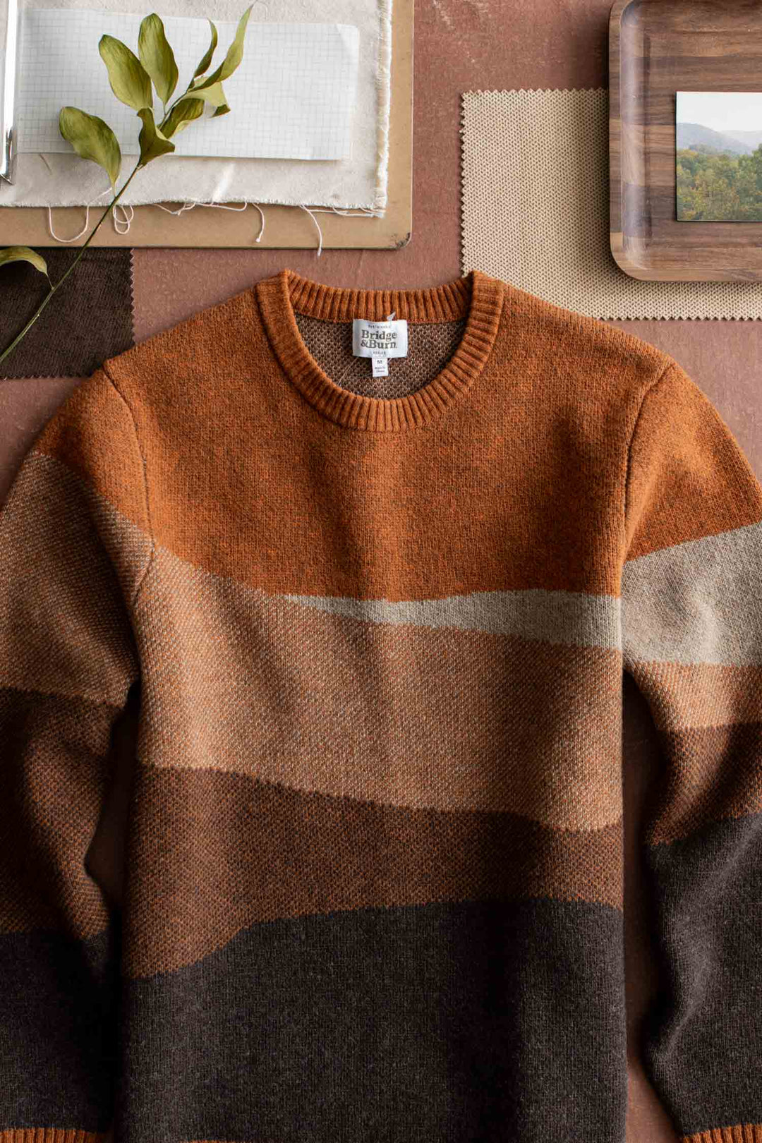 The Bridge & Burn Petra Sweater in Sandstone, featuring earthy horizontal stripes in brown, tan, and orange, is laid flat next to neutral fabric swatches, a wooden tray, and a leafy stem.