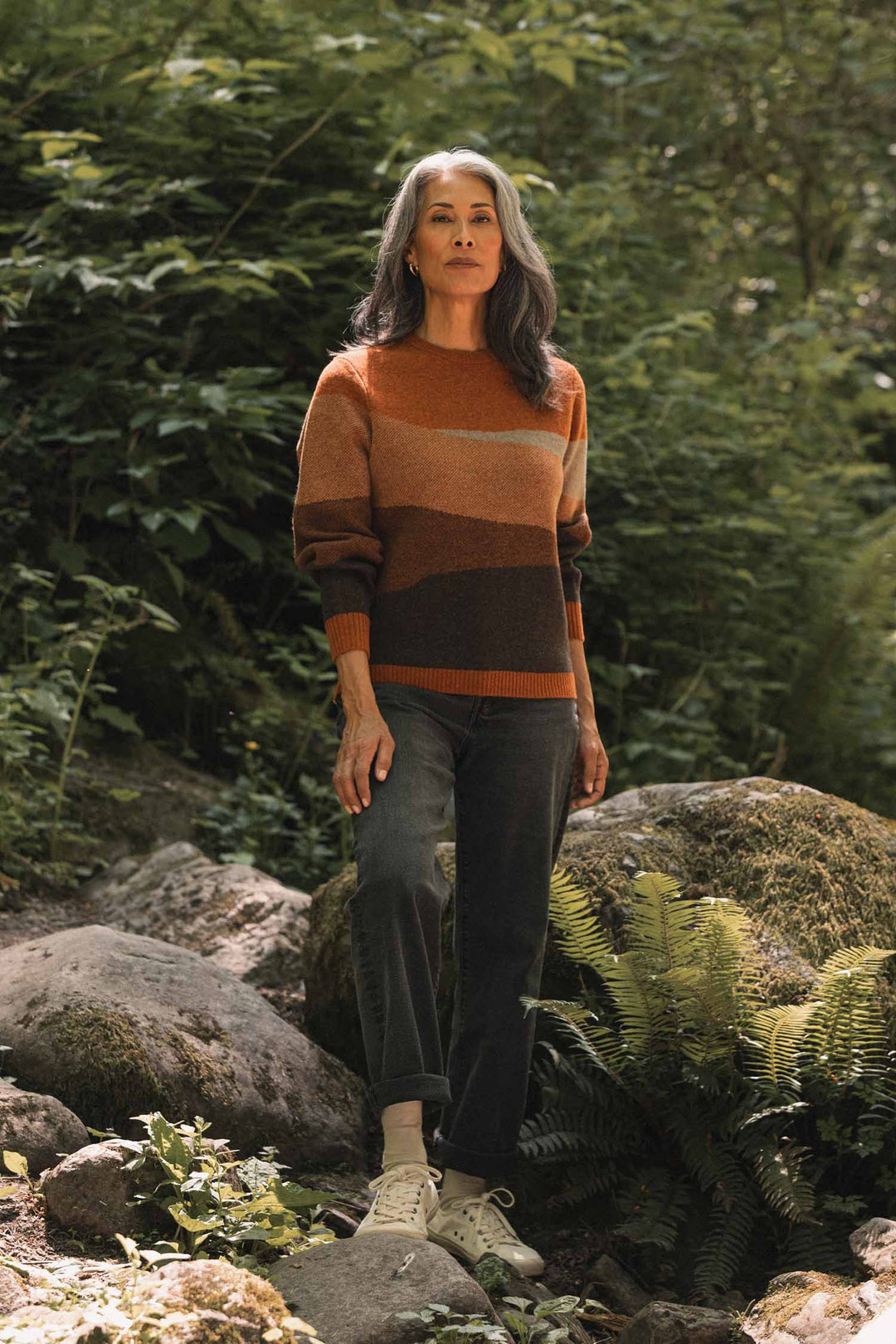 A woman with long gray hair stands on rocks in a green forest, wearing the Bridge & Burn Petra Sweater in Sandstone, dark jeans, and white sneakers. She gazes calmly ahead, surrounded by nature.