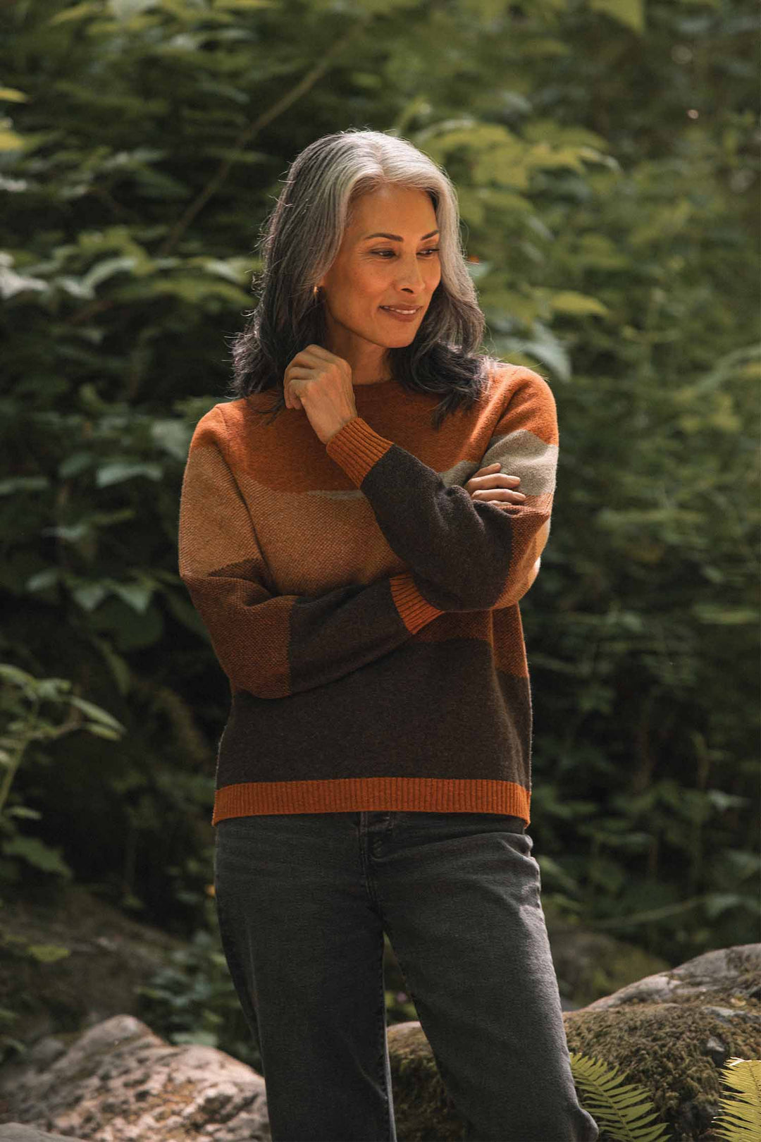 A woman with shoulder-length gray and black hair smiles outdoors in front of green foliage, arms crossed, wearing Bridge & Burn’s Petra Sweater in Sandstone—crafted from soft RWS-certified wool—and black jeans.