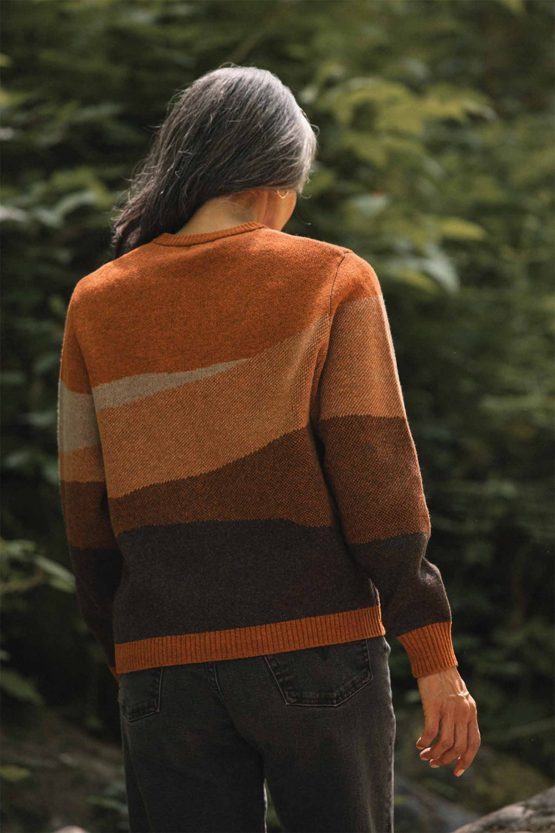 A person with long gray hair, wearing the Bridge & Burn Petra Sweater in Sandstone and dark jeans, stands outdoors facing away, surrounded by green foliage.