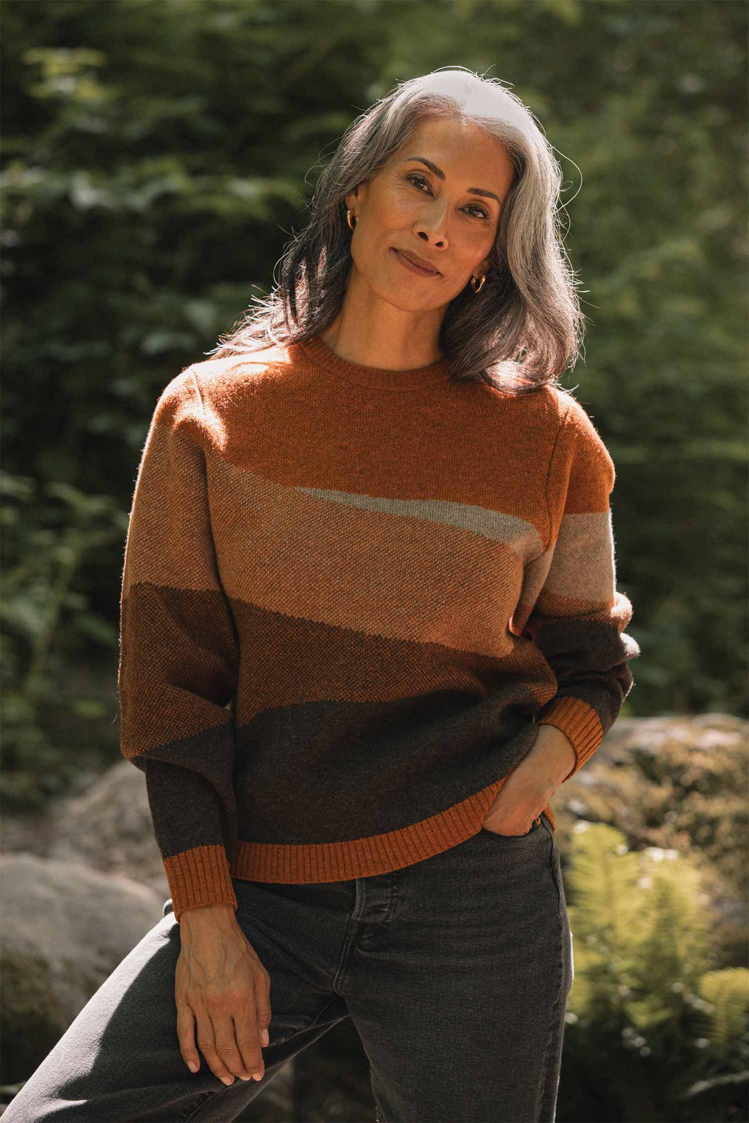 A woman with long gray hair smiles softly outdoors in natural sunlight, wearing the Bridge & Burn Petra Sweater in Sandstone—an earthy striped piece made from RWS-certified wool—with dark jeans, her hand casually in her pocket amid greenery and rocks.