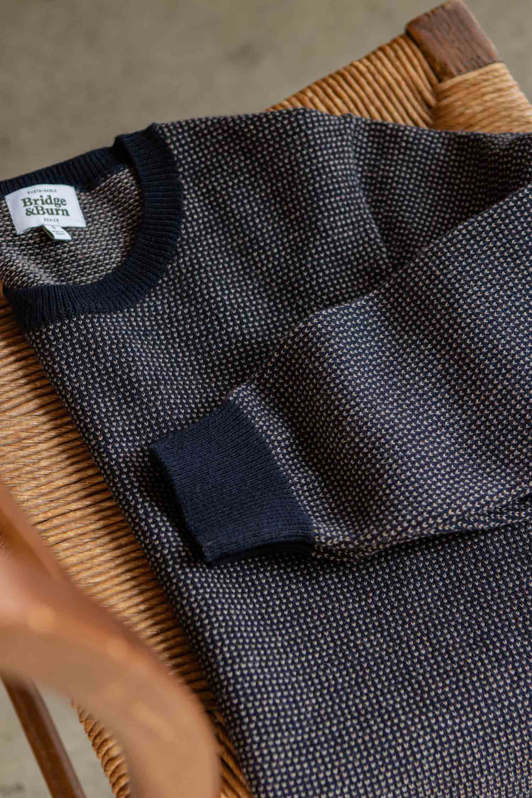 A Pickering Sweater in the Harbor Mist color by Bridge & Burn is neatly folded and set on a woven wooden chair. Made from soft Merino wool that meets the Responsible Wool Standard, this sweater features a subtle texture along with ribbed cuffs and collar.