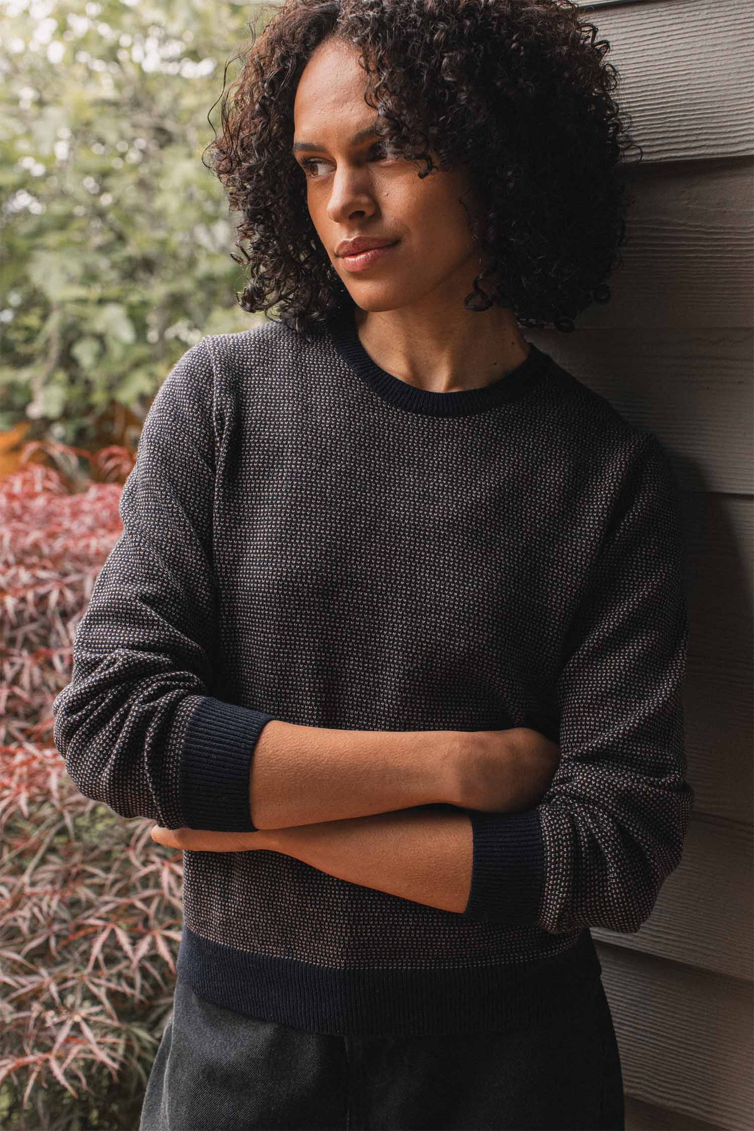 A person with curly hair stands by a wall, arms crossed, wearing the Bridge & Burn Pickering Sweater in Harbor Mist. Green foliage and red leaves provide a backdrop that emphasizes the responsible craftsmanship of this Responsible Wool Standard garment.
