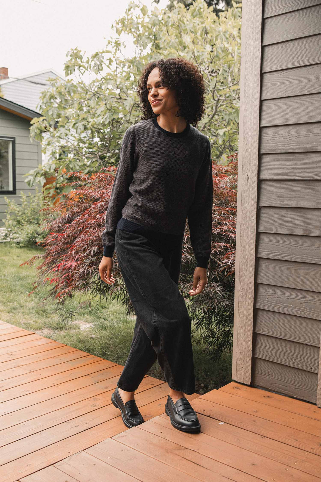 A woman with curly hair, dressed in a dark Harbor Mist Pickering Sweater by Bridge & Burn, black pants, and black loafers, stands on a wooden porch beside a gray house, surrounded by greenery and red-leafed plants.