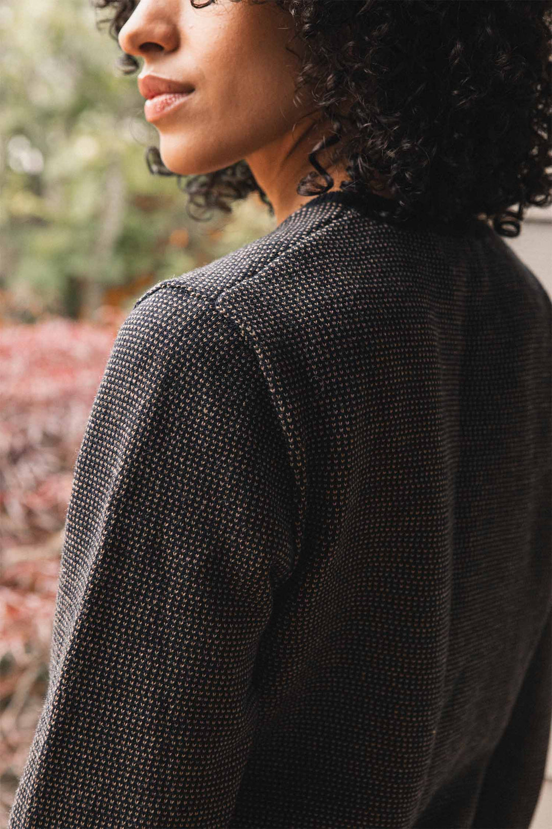 A person with curly hair is shown from the side, wearing a textured dark jacket layered over the Bridge & Burn Pickering Sweater in Harbor Mist. The softly blurred background features greenery and reddish plants.