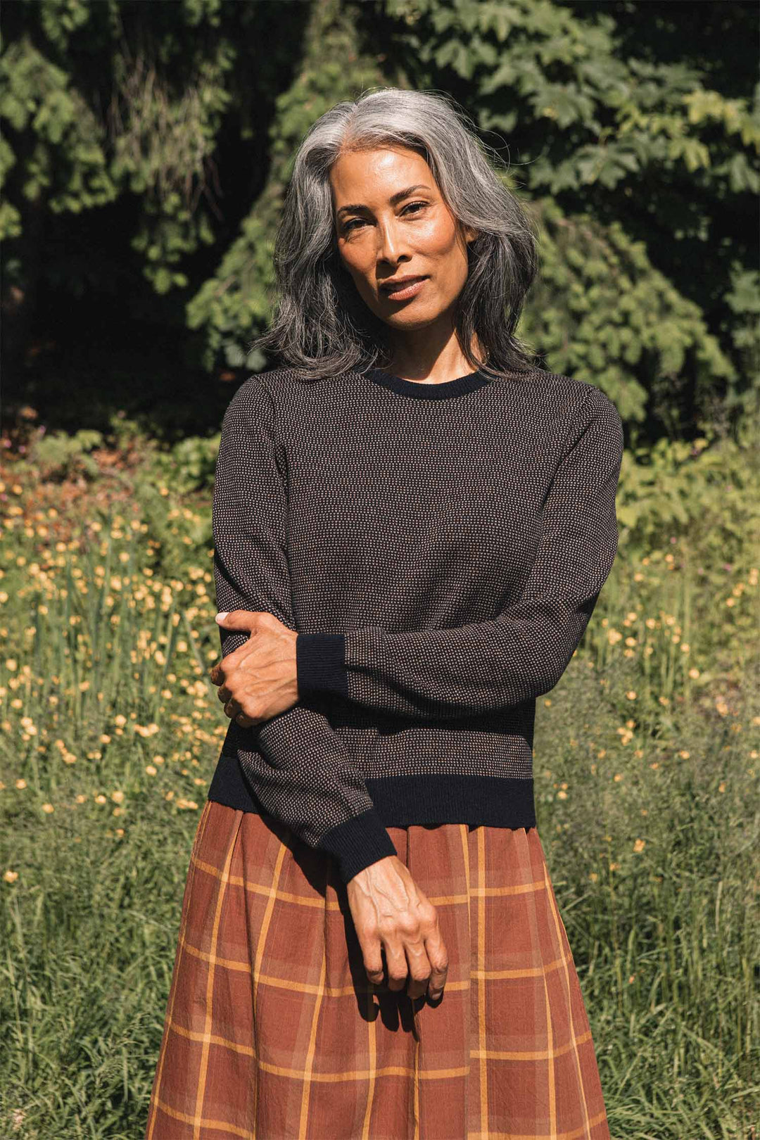 A woman with long gray hair stands outdoors, surrounded by greenery and yellow flowers, wearing the Bridge & Burn Pickering Sweater in Harbor Mist—a dark Merino wool piece made according to the Responsible Wool Standard for comfort and sustainability—paired with a brown plaid skirt.