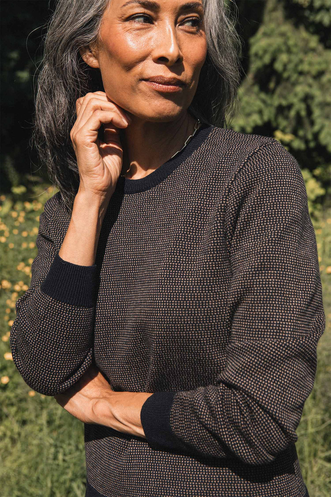 A person with long gray hair stands outdoors, surrounded by greenery and yellow flowers, wearing the Bridge & Burn Pickering Sweater in Harbor Mist—a textured dark Merino wool sweater that meets the Responsible Wool Standard—with one hand touching their chin and the other arm crossed.