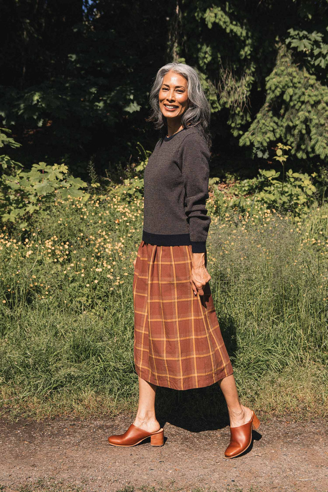 A woman with long gray hair walks outside on a path, smiling as she passes green foliage and yellow flowers. She is wearing the Bridge & Burn Pickering Sweater in Harbor Mist, crafted from Responsible Wool Standard-certified Merino wool, along with a brown plaid skirt and brown heeled shoes.