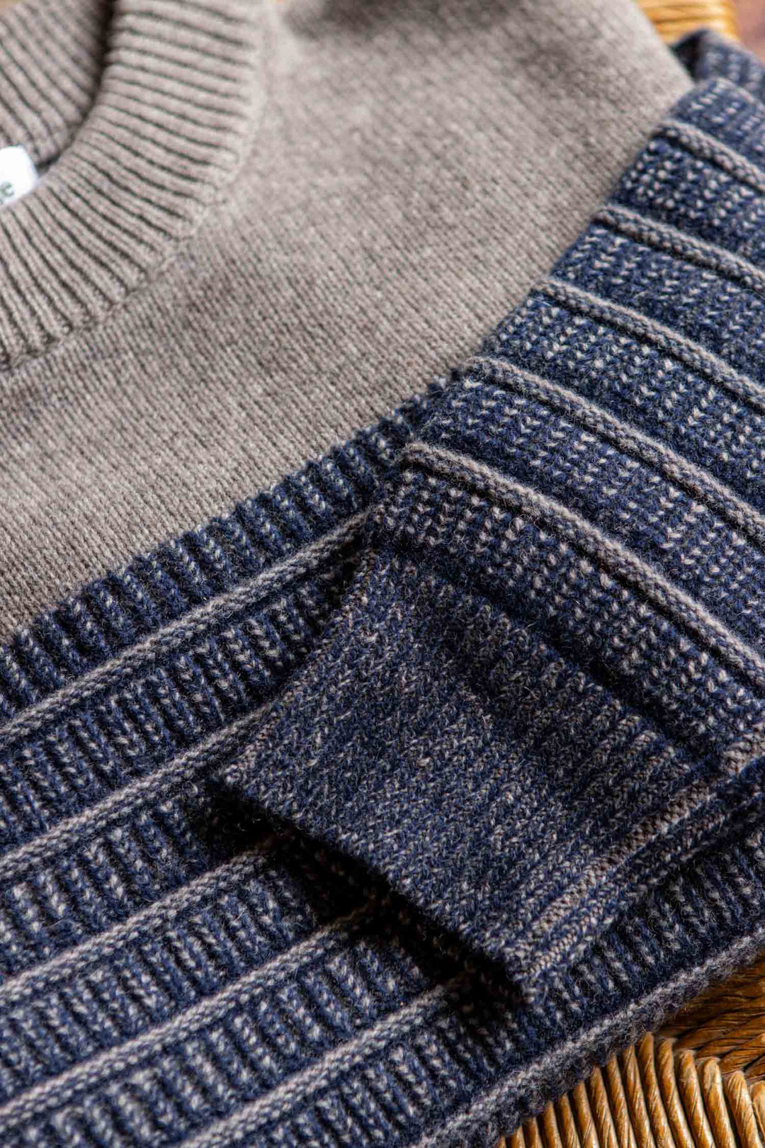 A close-up of the Bridge & Burn Sabine Sweater in Mariner Stripe, showcasing its folded heavyweight knit with a solid taupe upper and dark blue and gray striped pattern on the sleeves and lower body, resting on a wicker chair.