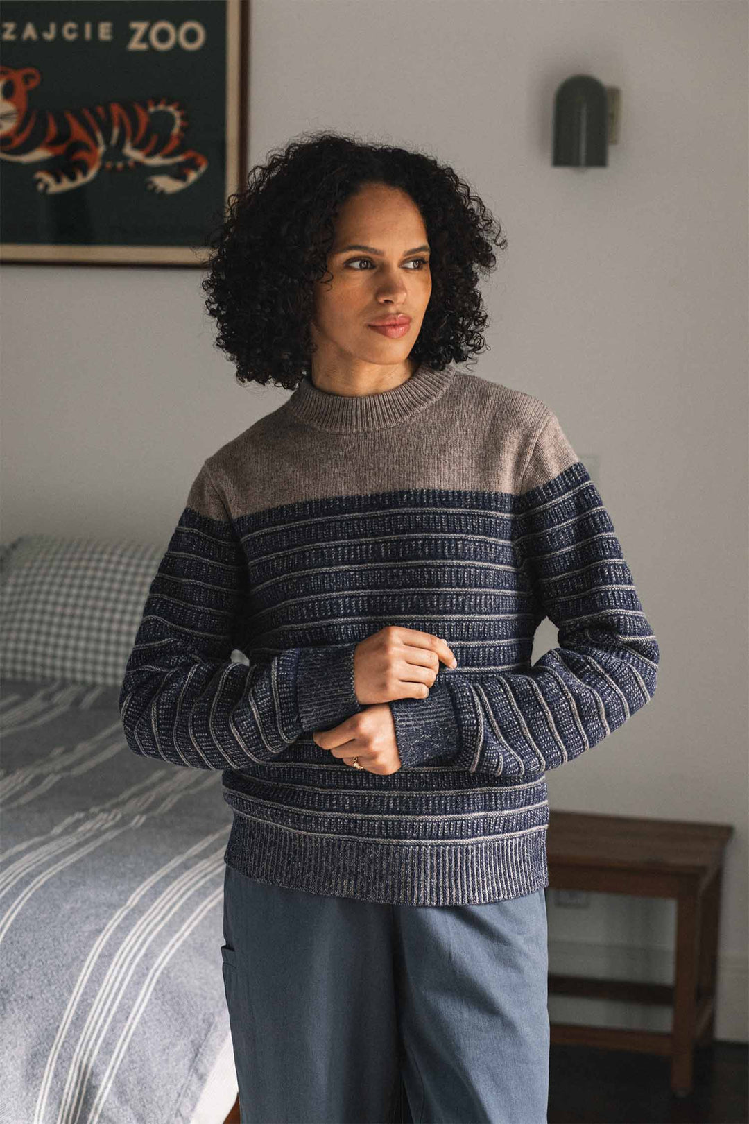 A person with curly hair stands indoors wearing the Bridge & Burn Sabine Sweater in Mariner Stripe and blue pants. Behind them is a bed with checkered bedding, a wooden nightstand, and a framed poster of a tiger on the wall.