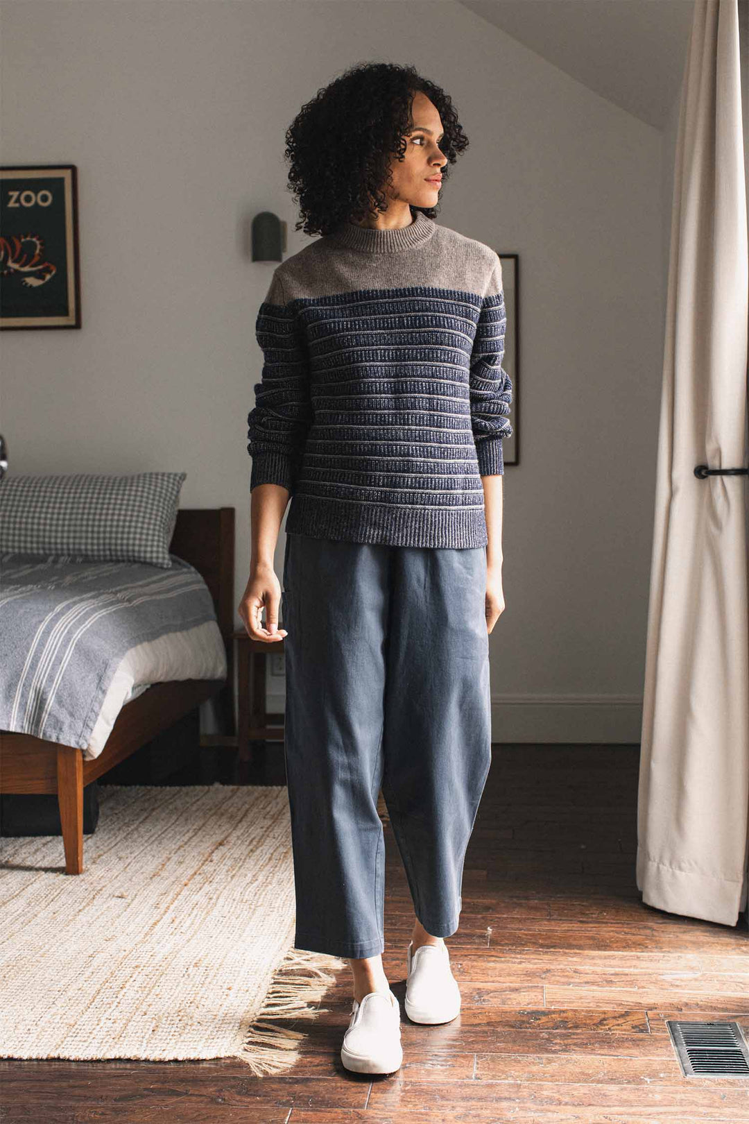 A person with curly hair, wearing the Bridge & Burn Sabine Sweater in Mariner Stripe, loose blue pants, and white shoes stands in a bedroom with a bed, rug, and framed art on the wall.