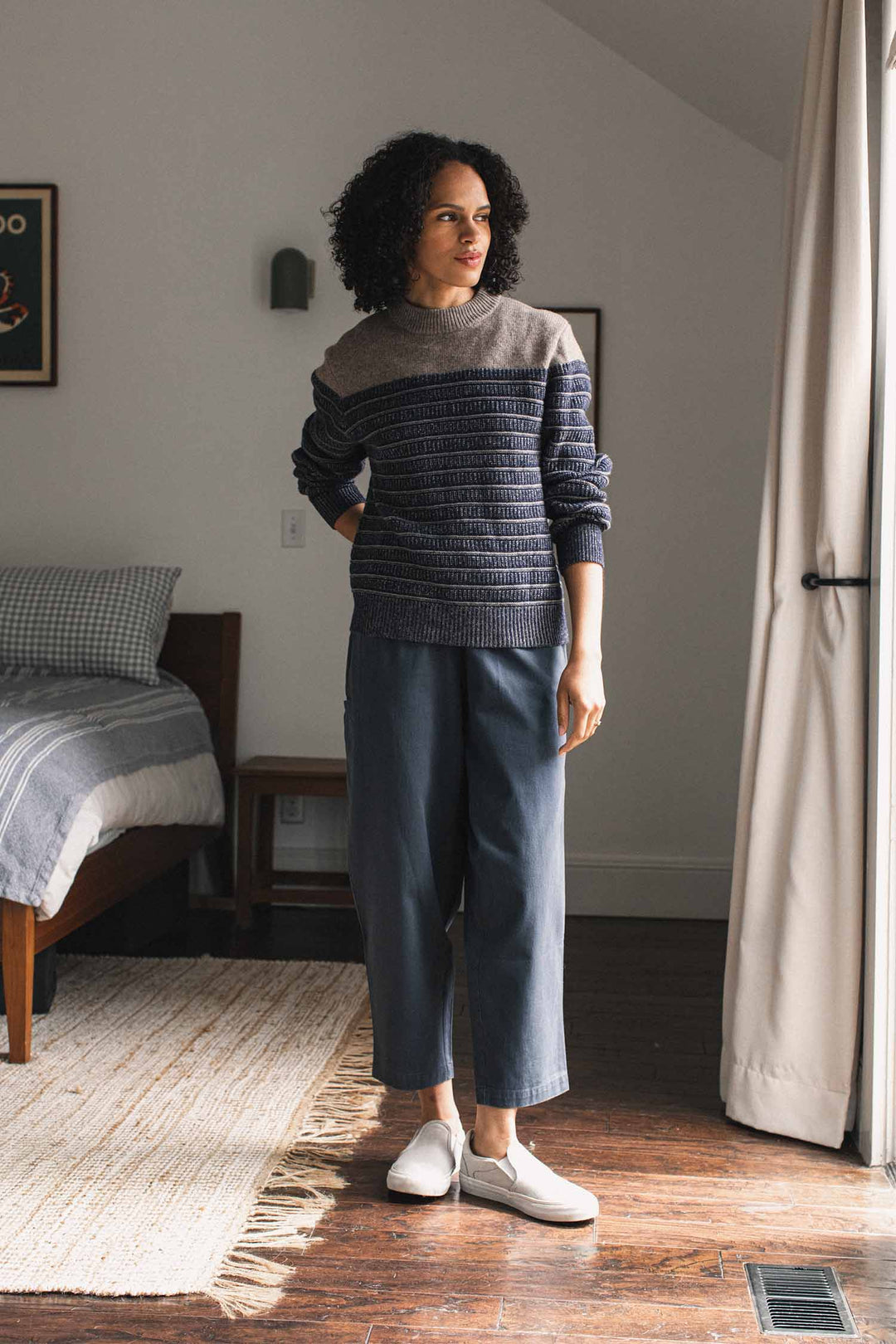 A woman with curly hair stands indoors near a bed, wearing the Bridge & Burn Sabine Sweater in Mariner Stripe, along with wide-legged blue pants and white shoes. She gazes to the side as natural light streams through a nearby glass door.