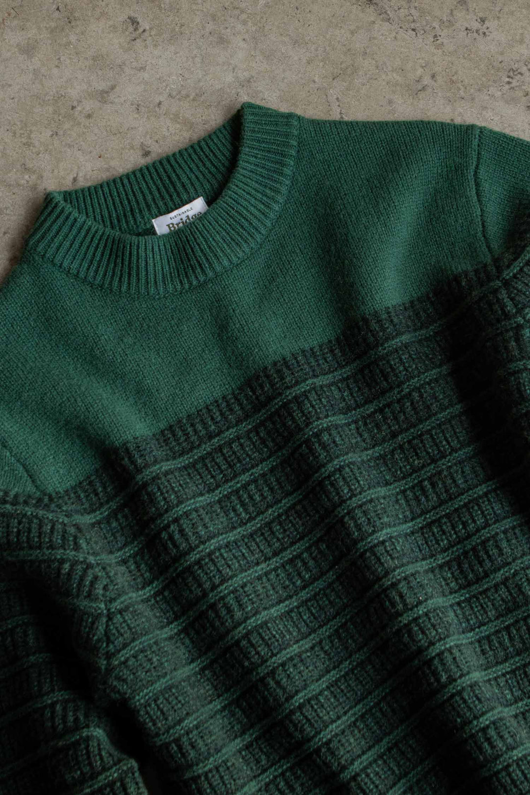 The Sabine Sweater in Pine Shadow by Bridge & Burn is a heavyweight green sweater featuring a ribbed collar and a textured horizontal pattern across the body, shown laid flat on a rough, beige surface.