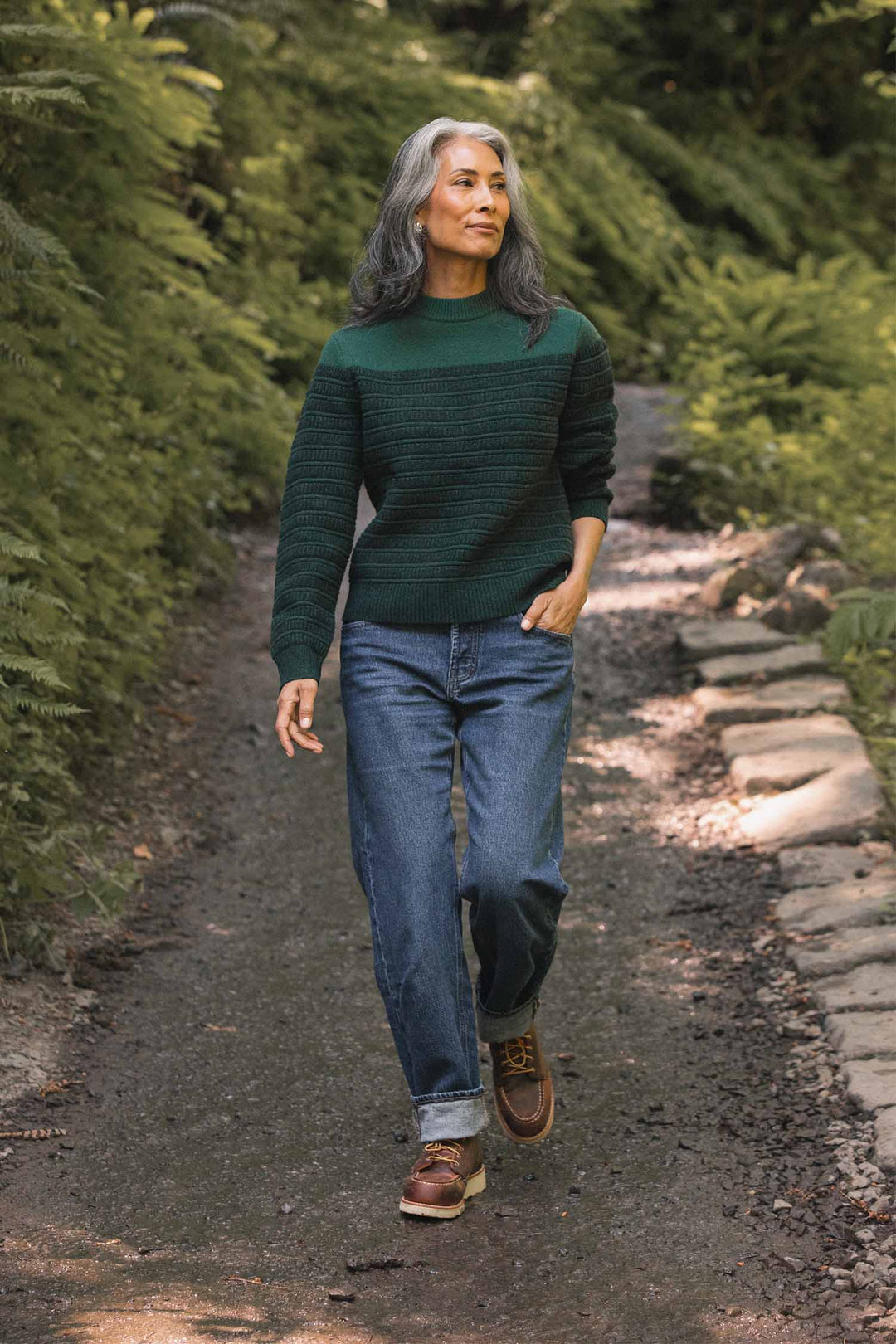 A woman with long gray hair walks on a leafy path, surrounded by lush greenery. She wears the Bridge & Burn Sabine Sweater in Pine Shadow, paired with blue jeans and brown shoes, with one hand in her pocket.