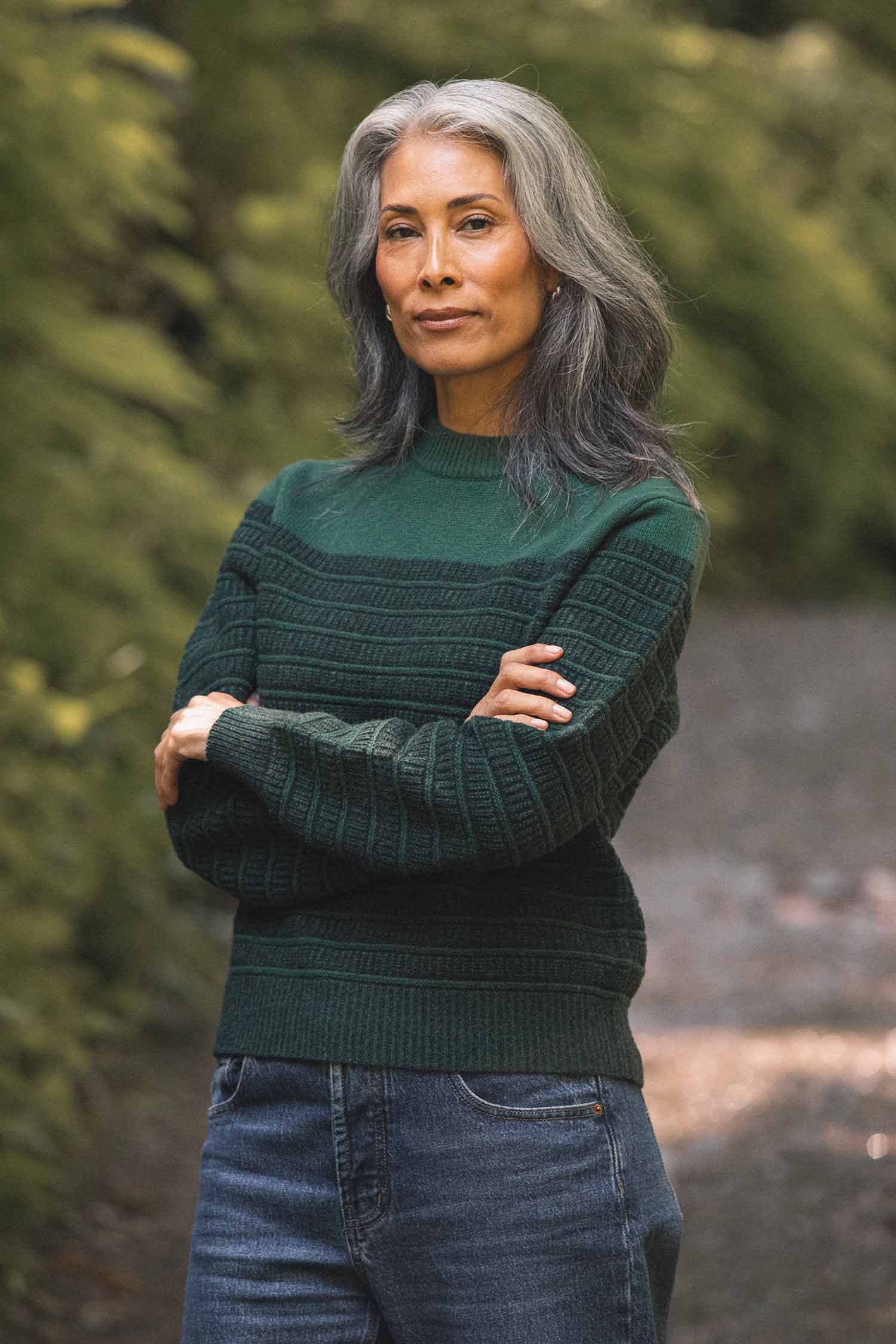 A woman with long gray hair stands outdoors on a path, wearing the Bridge & Burn Sabine Sweater in Pine Shadow and blue jeans. She has her arms crossed and looks confidently at the camera, surrounded by greenery.