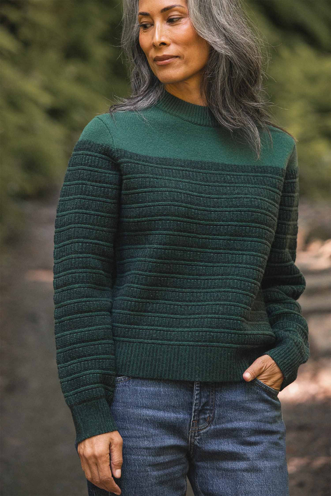 A woman with long gray hair stands outdoors, one hand in her pocket, wearing the Bridge & Burn Sabine Sweater in Pine Shadow and blue jeans. Blurred greenery forms the background.