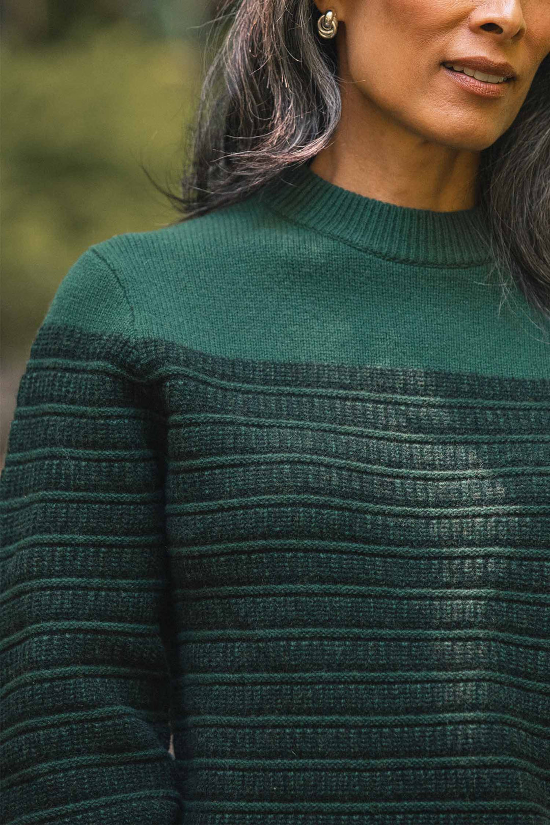 A woman with long dark hair, wearing gold stud earrings and the Bridge & Burn Sabine Sweater in Pine Shadow, stands outdoors against a softly blurred natural background.