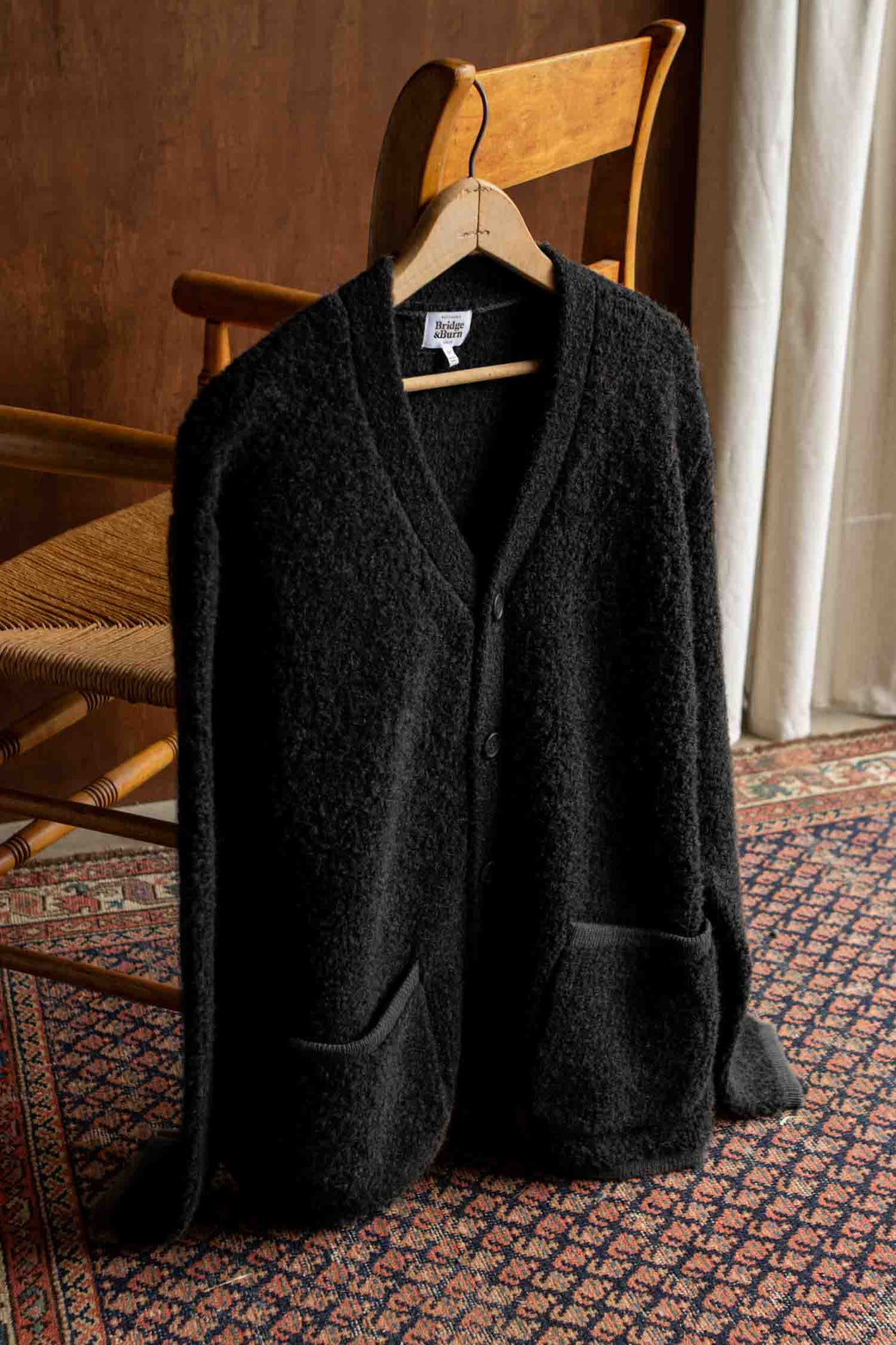 A Bridge & Burn Whit Cardigan in black, featuring a textured knit and two front pockets, hangs on a wooden hanger draped over the back of a wooden chair, set atop a patterned rug near a window with a curtain.