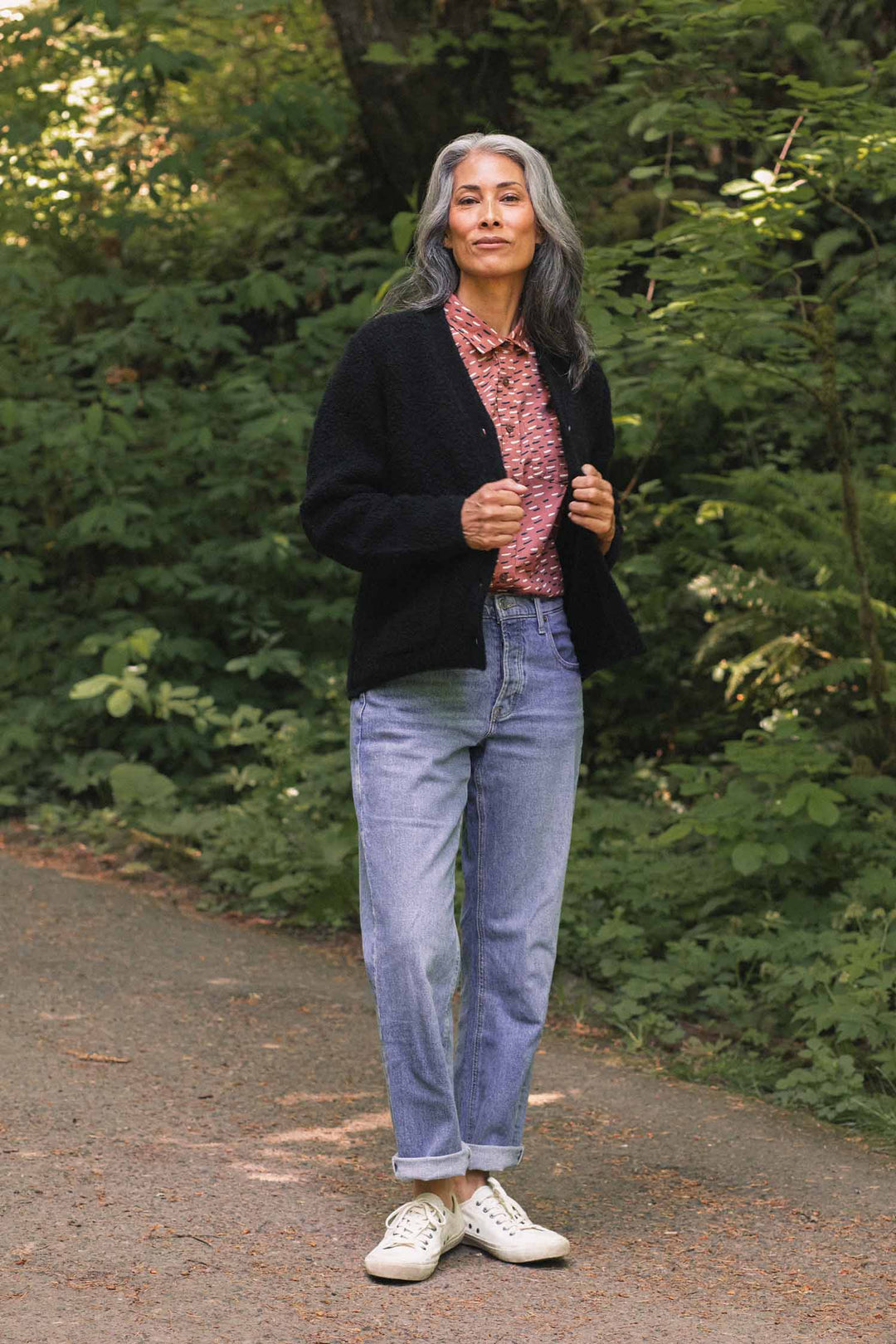 A woman with long gray hair stands on a path in a wooded area, wearing the Bridge & Burn Whit Cardigan in black over a patterned V-neck shirt, light blue jeans, and white sneakers. She looks at the camera with a relaxed expression.