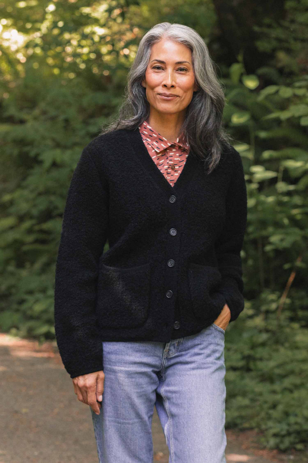 A woman with long gray hair stands outdoors, smiling gently. She wears the Bridge & Burn Whit Cardigan in black—a textured knit—layered over a red patterned V-neck shirt and blue jeans, with one hand in her pocket. Green foliage and a wooded path are visible in the background.