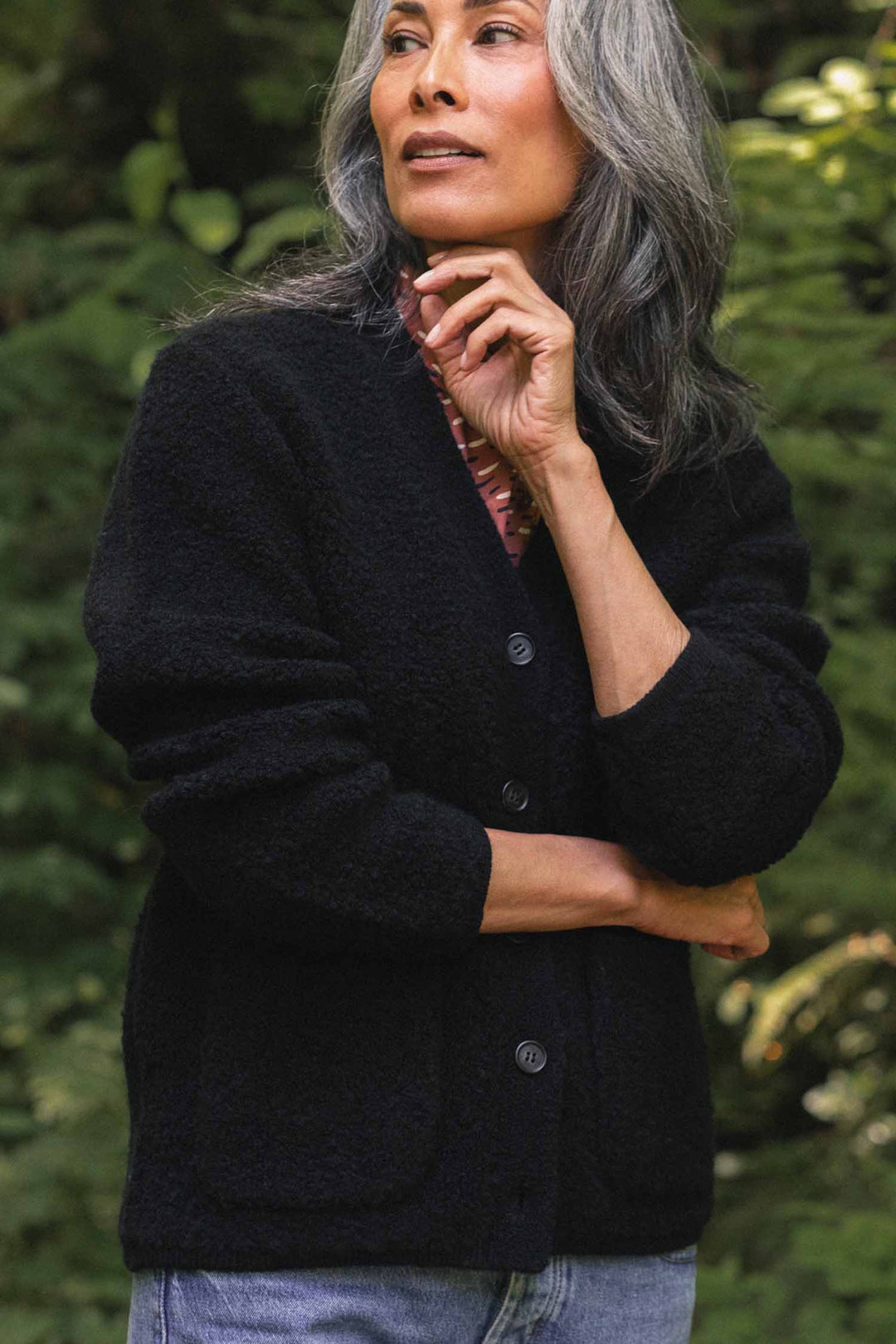 A person with long gray hair stands outdoors surrounded by greenery, wearing the Bridge & Burn Whit Cardigan in black—a textured knit, V-neck style—paired with light jeans. One hand rests thoughtfully on their chin while the other is crossed over their body.