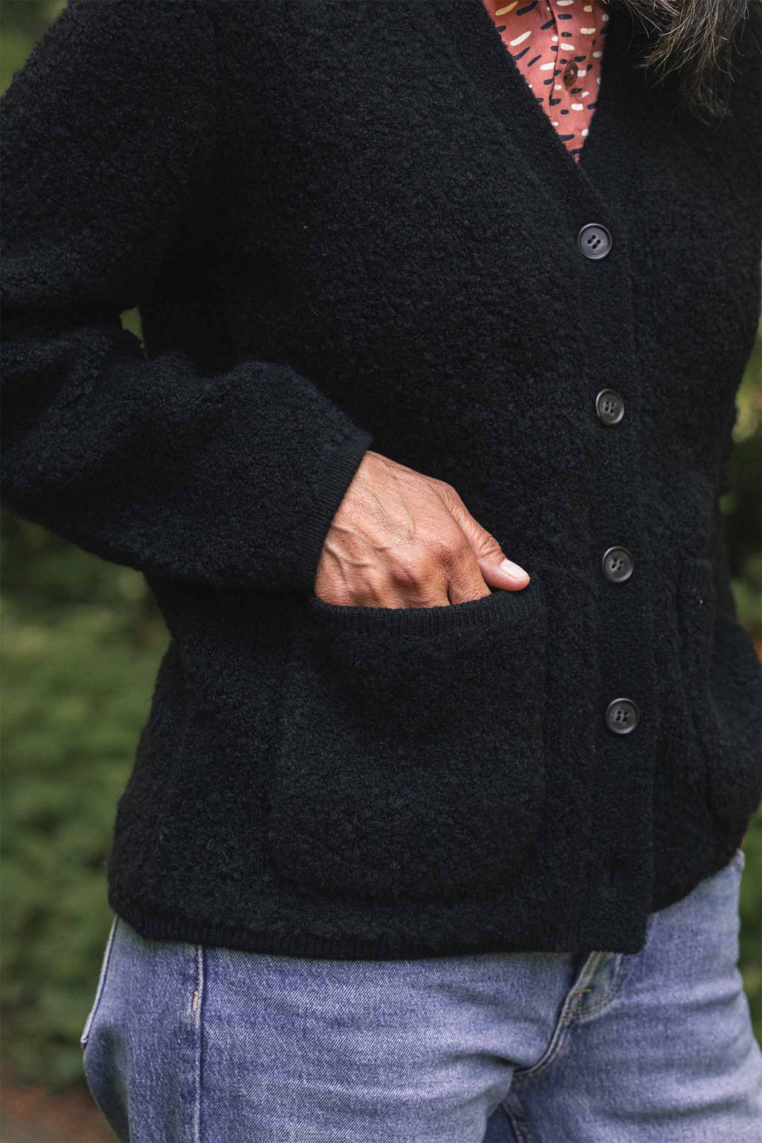 A person stands outdoors with one hand in a pocket, wearing the Bridge & Burn Whit Cardigan in black over a pink patterned shirt and blue jeans.