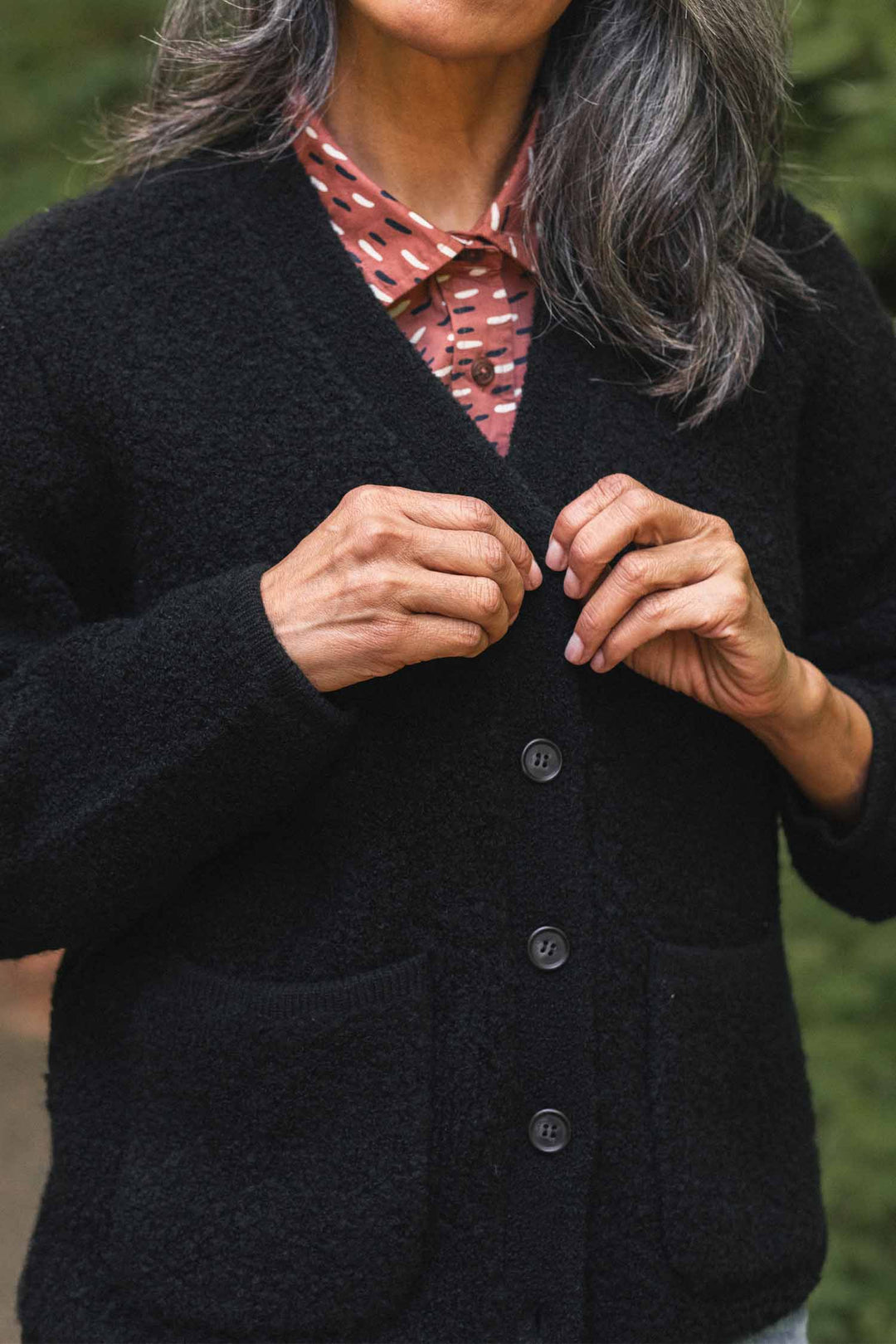 A person with long gray hair buttons the Bridge & Burn Whit Cardigan in black—a textured knit V-neck style—over a patterned pink shirt. The image, taken outdoors, focuses on their hands and upper torso.