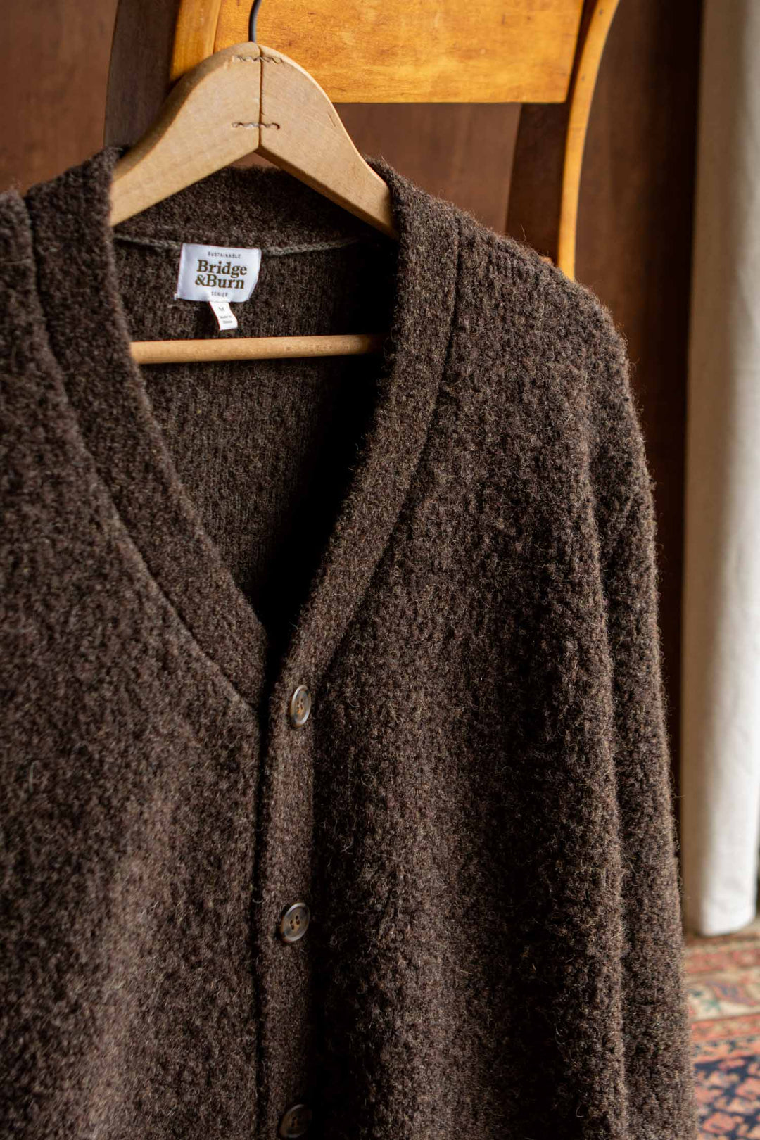 A textured knit dark olive V-neck cardigan with patch pockets and buttons hangs on a wooden hanger over the back of a wooden chair. The label on the sweater reads Bridge & Burn, and the style is called Whit Cardigan.