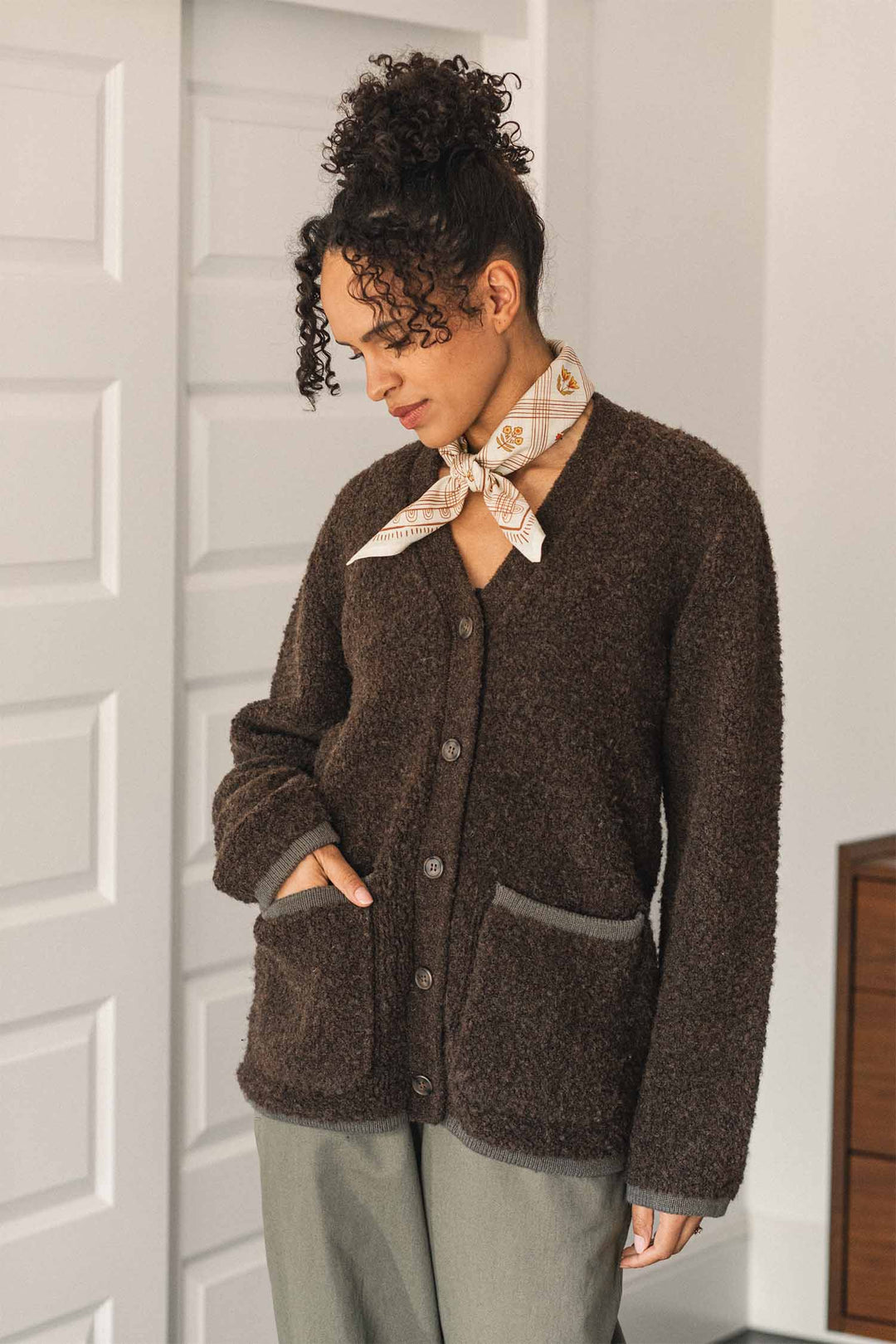 A woman stands indoors near a white paneled door, wearing the Bridge & Burn Whit Cardigan in Dark Olive—a cozy V-neck cardigan with gray patch pockets and textured knit details—paired with olive pants and a cream patterned scarf tied around her neck.