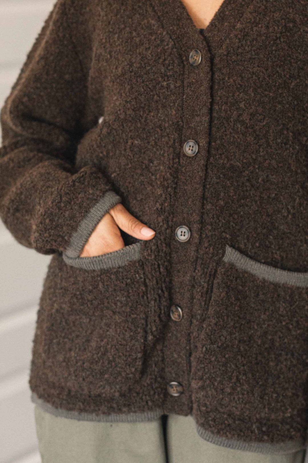 A person wearing the Bridge & Burn Whit Cardigan in Dark Olive—a textured knit V-neck style featuring patch pockets and gray trim—has one hand in a pocket. The person's face is not visible.