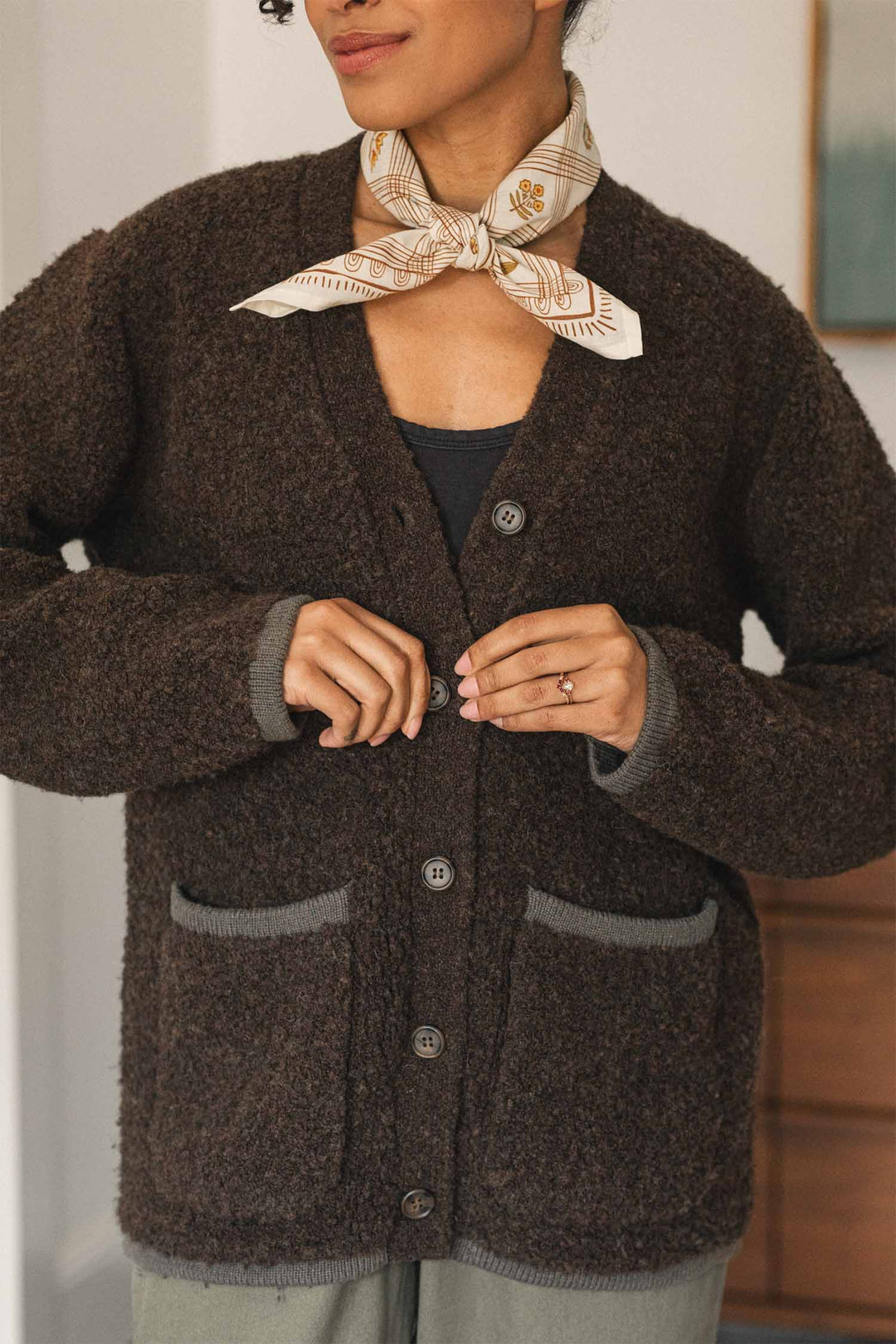 A woman wearing the Bridge & Burn Whit Cardigan in dark olive, featuring textured knit fabric and patch pockets with gray trim, stands indoors with a light printed scarf around her neck, a black top, and an engagement ring on her finger, partially smiling.