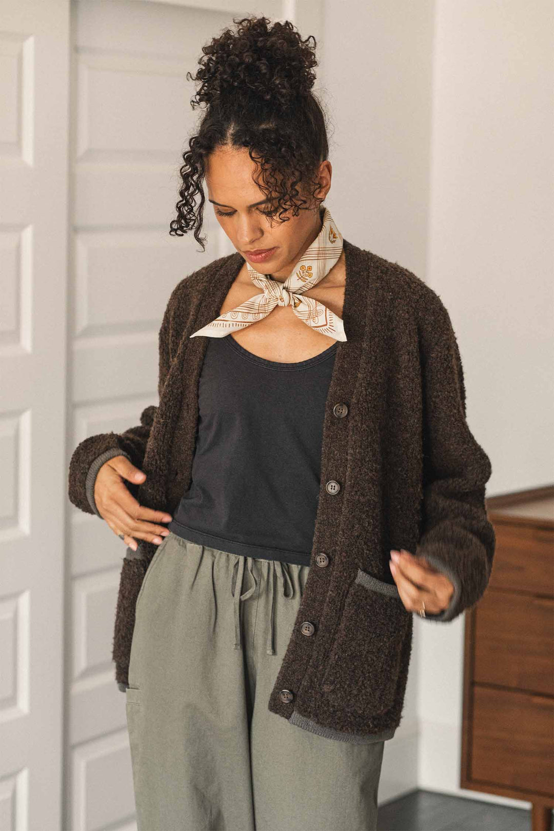 A woman with curly hair in a bun stands indoors wearing the Bridge & Burn Whit Cardigan in dark olive—a textured knit V-neck cardigan with patch pockets—layered over a black top and gray pants, accessorized with a light-colored scarf tied around her neck.