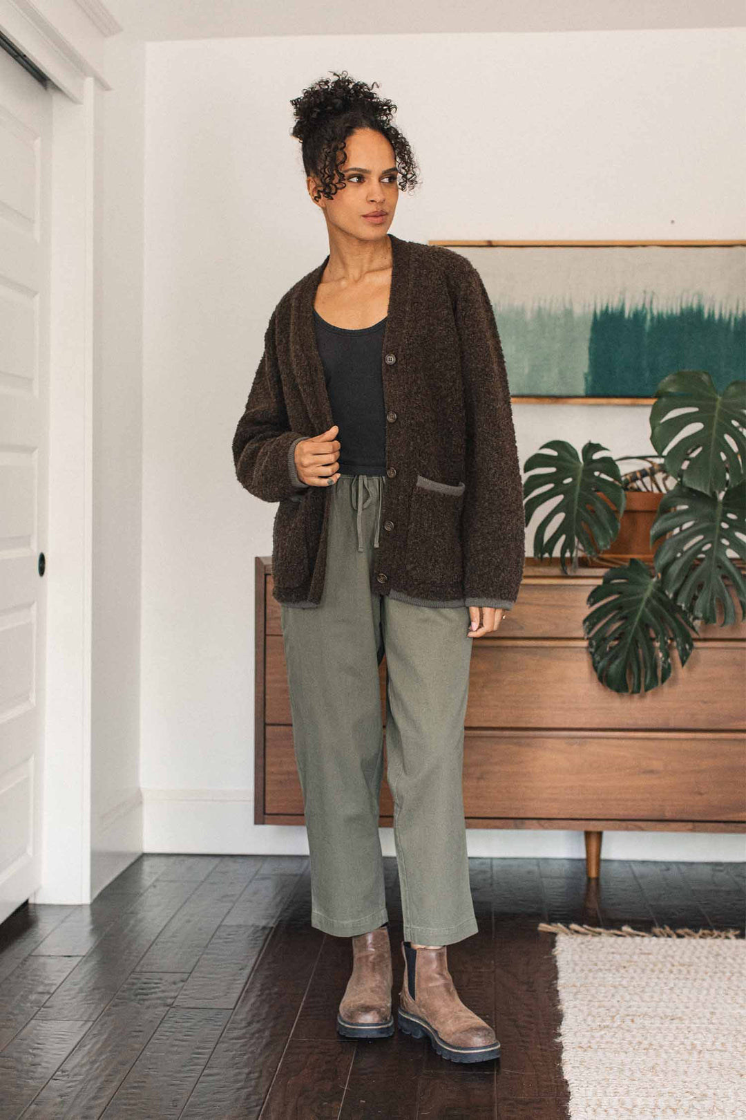 A woman stands indoors wearing the Bridge & Burn Whit Cardigan in Dark Olive over a black top, paired with olive green pants and brown boots. A houseplant and framed art are visible in the background.