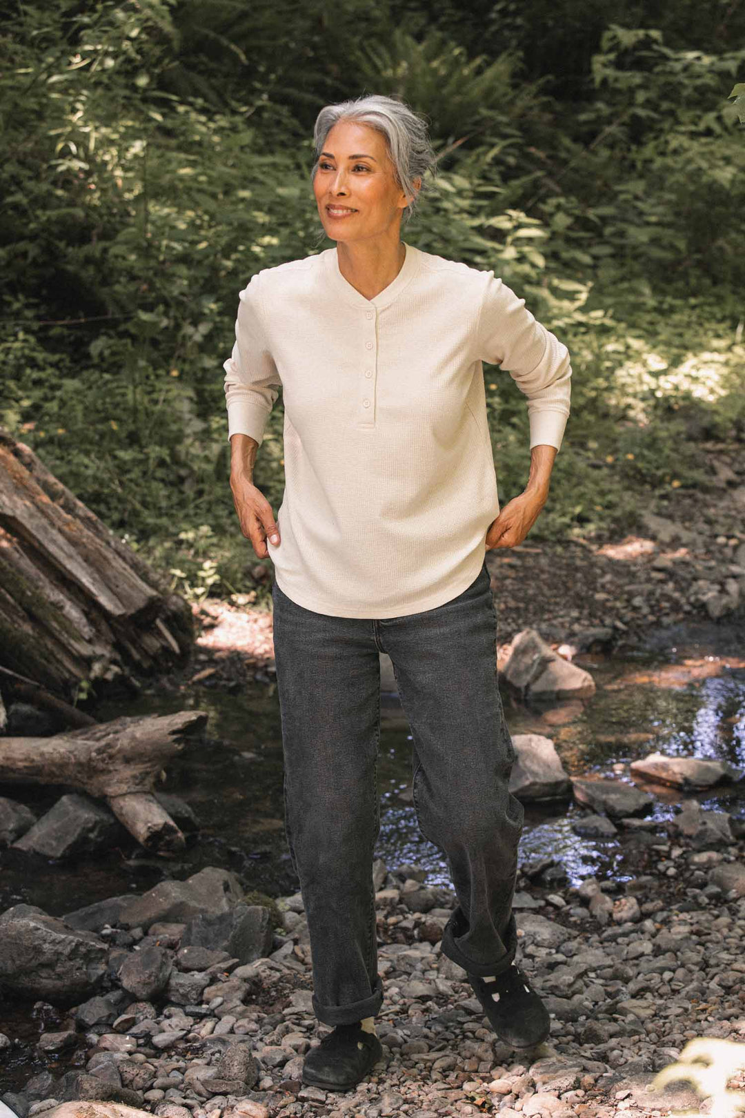 A person with gray hair, wearing the Bridge & Burn Women’s Recycled Waffle Henley in Ivory and dark pants, stands on a rocky path beside a small stream in a lush, wooded area.