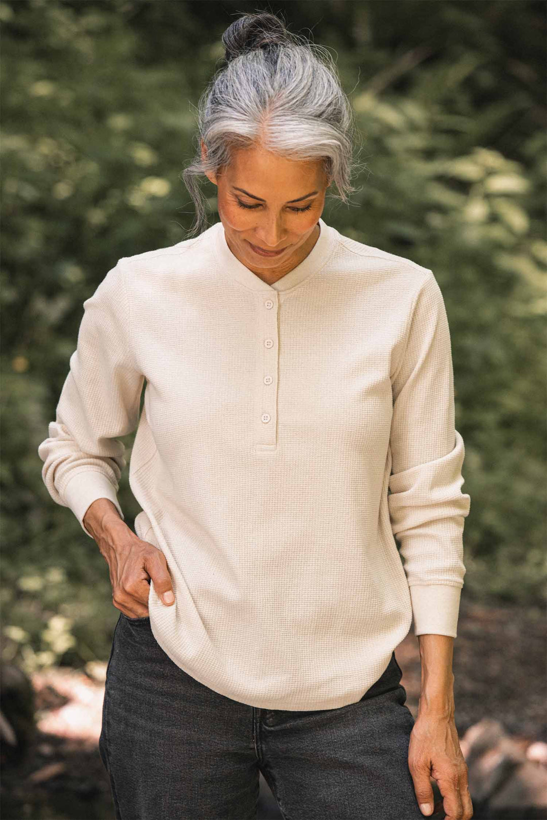 A woman with gray hair in a bun looks down while wearing the Bridge & Burn Women’s Recycled Waffle Henley in Ivory and dark pants, standing outdoors with green foliage in the background.