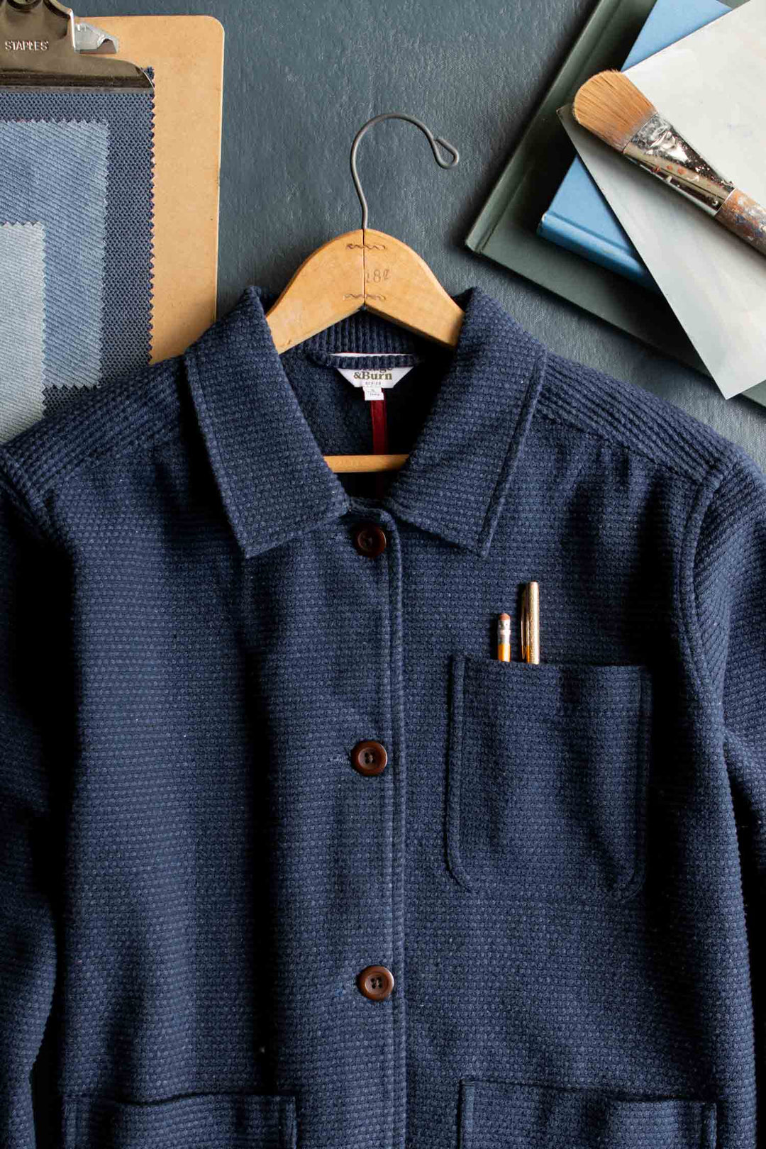 A Women’s Studio Chore Coat in Indigo by Bridge & Burn, crafted from organic cotton with a textured finish, hangs on a hanger with pens tucked into the chest pocket, displayed alongside swatches, a clipboard, books, and a paintbrush on a dark surface.