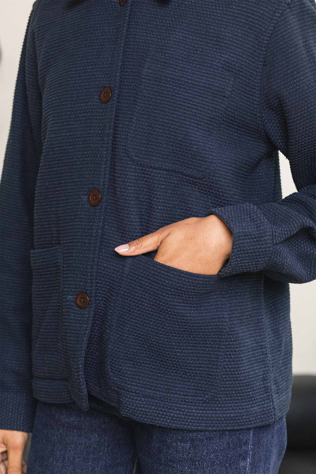 A person wearing the Bridge & Burn Women’s Studio Chore Coat in Indigo—a dark blue textured organic cotton workwear jacket with brown buttons—paired with jeans, with one hand in the jacket pocket.