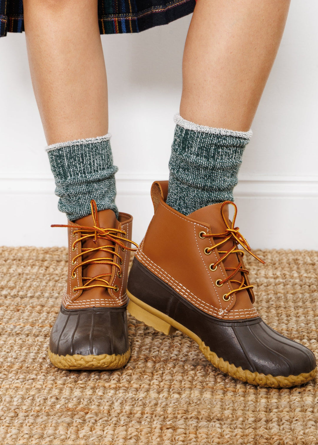 Someone wearing American Trench - Sunday Sock in Bottle Green stands on a woven beige rug in brown duck boots, with a plaid skirt visible above the knees.