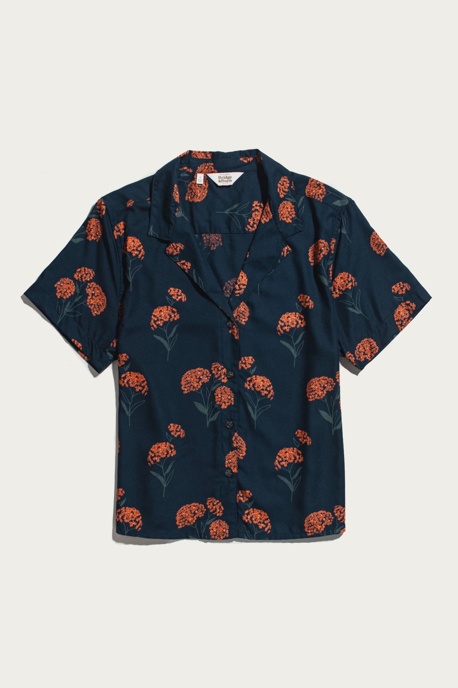 Bridge & Burn's Mila Cropped Shirt in the Wildflower print—a short-sleeve, collared navy button-up crafted from modal and adorned with a scattered pattern of red-orange flowers and green stems—is shown laid flat on a white background.
