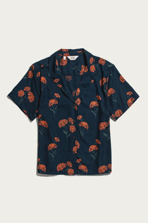 Bridge & Burn's Mila Cropped Shirt in the Wildflower print—a short-sleeve, collared navy button-up crafted from modal and adorned with a scattered pattern of red-orange flowers and green stems—is shown laid flat on a white background.