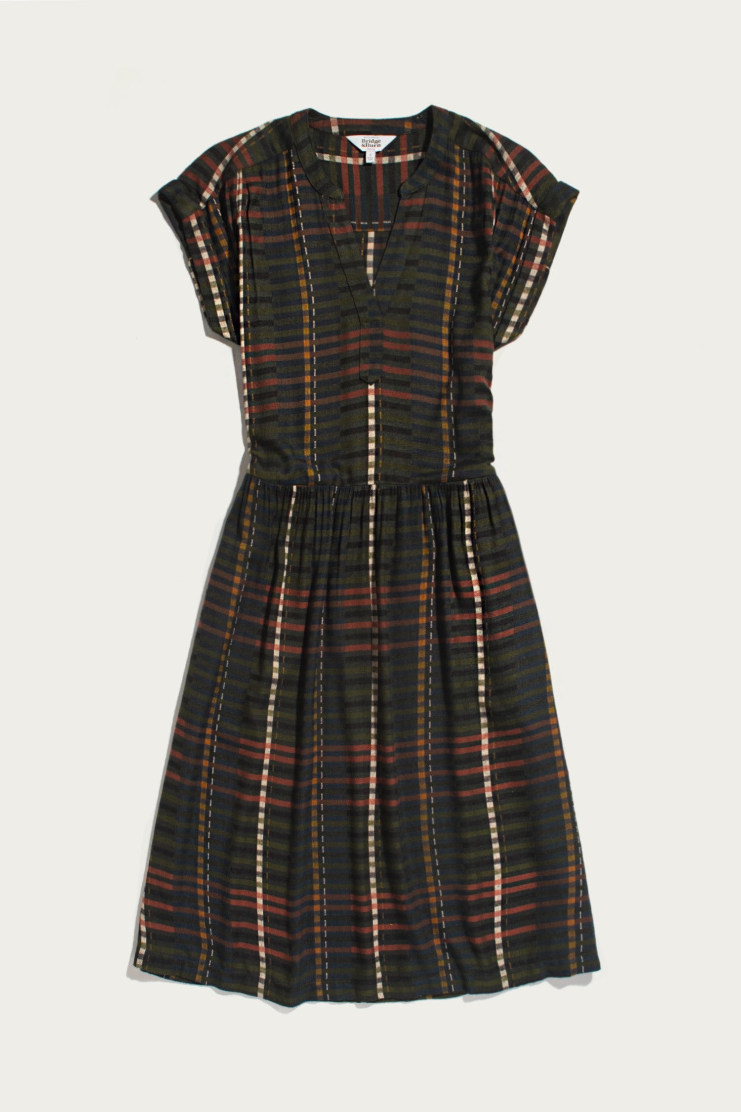 The Bridge & Burn Ira Dress in Olive Stripe is a short-sleeve, knee-length pullover dress with a dark multicolored striped pattern, featuring a gathered waist and split neckline. Crafted from a soft modal-linen blend, it is shown laid flat on a white background.