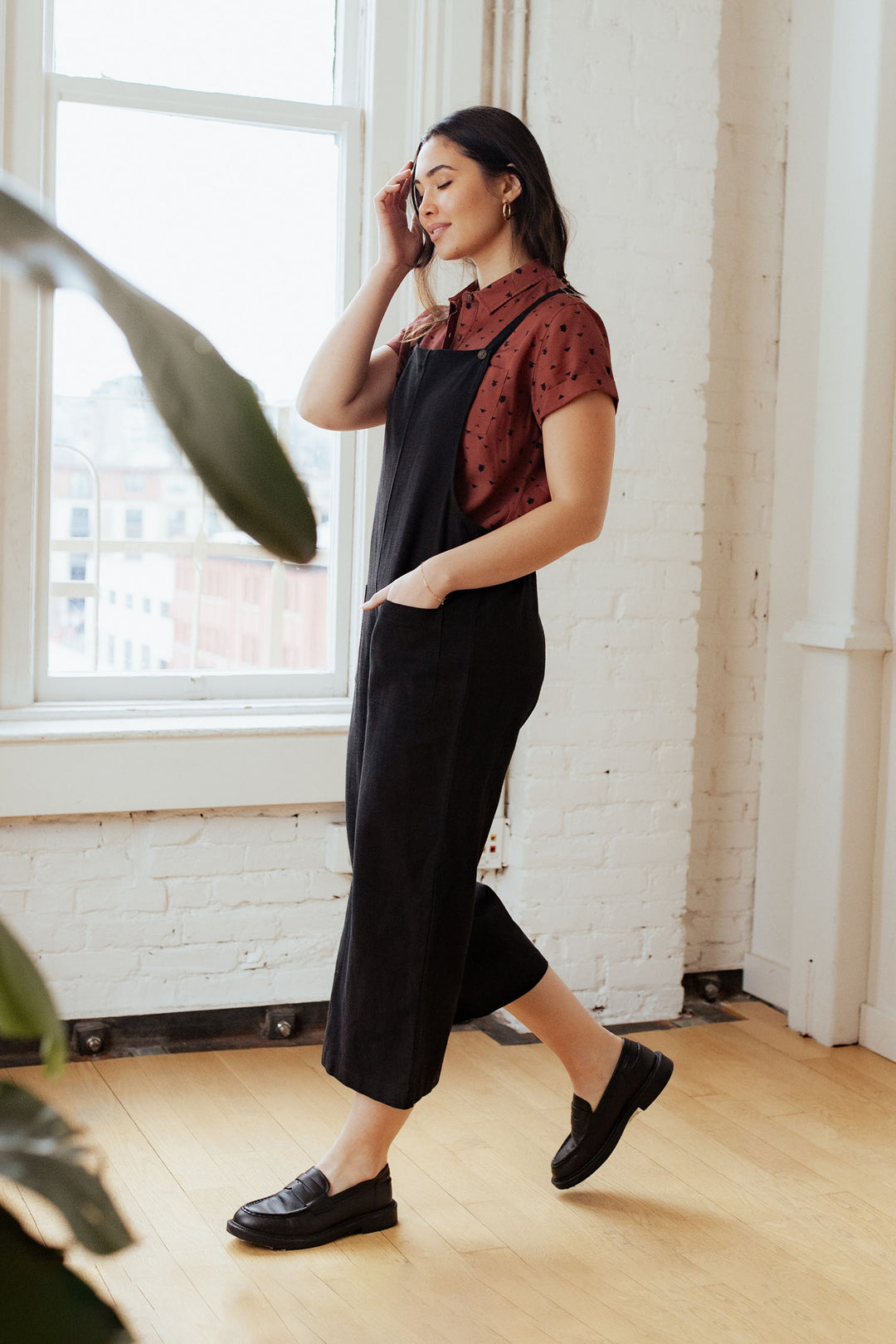 A woman stands indoors near large windows, wearing the Bridge & Burn Edie Overall in Black styled over a maroon blouse and paired with black loafers, with one hand in her pocket and the other touching her hair.