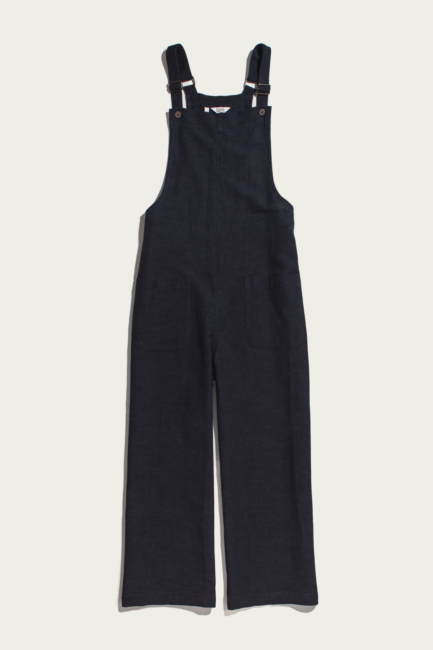 The Edie Overall in Navy by Bridge & Burn is laid flat on a white background, featuring adjustable straps and front patch pockets. Designed for a relaxed fit, these dark blue denim overalls blend comfort with timeless style.
