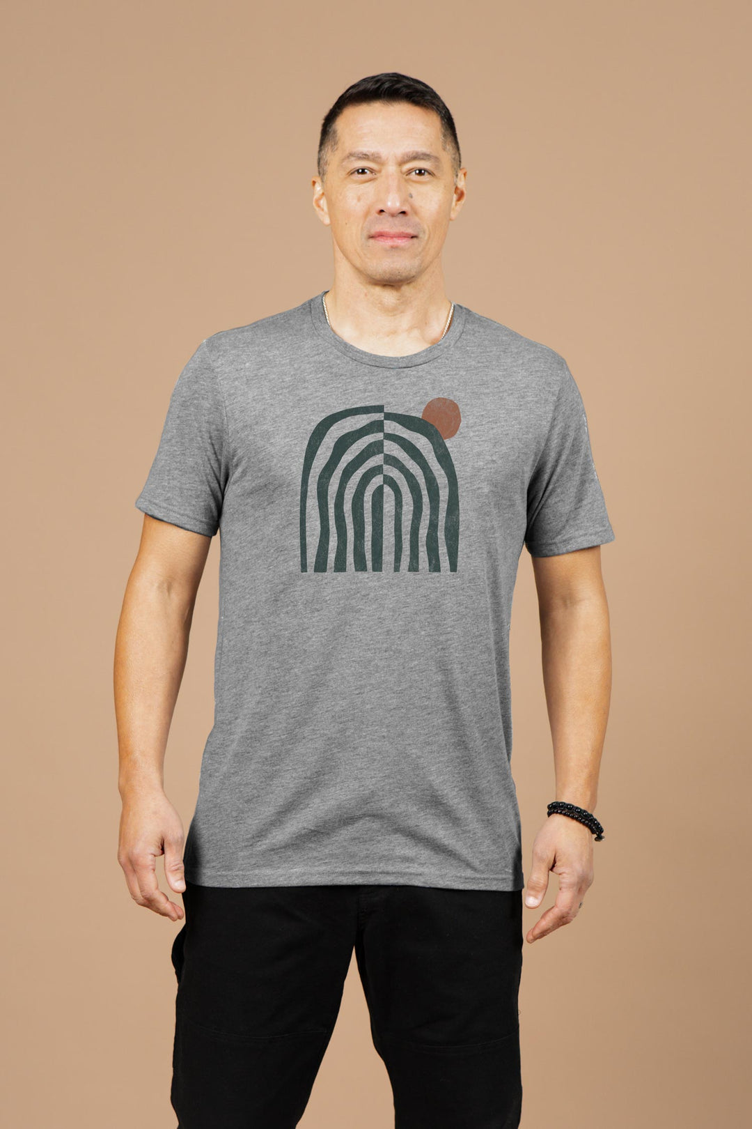 A man stands against a beige background, wearing the Bridge & Burn Men's Canopy Tee in grey—an organic cotton shirt featuring an abstract green arch with a brown circle—paired with black pants and a black beaded bracelet.