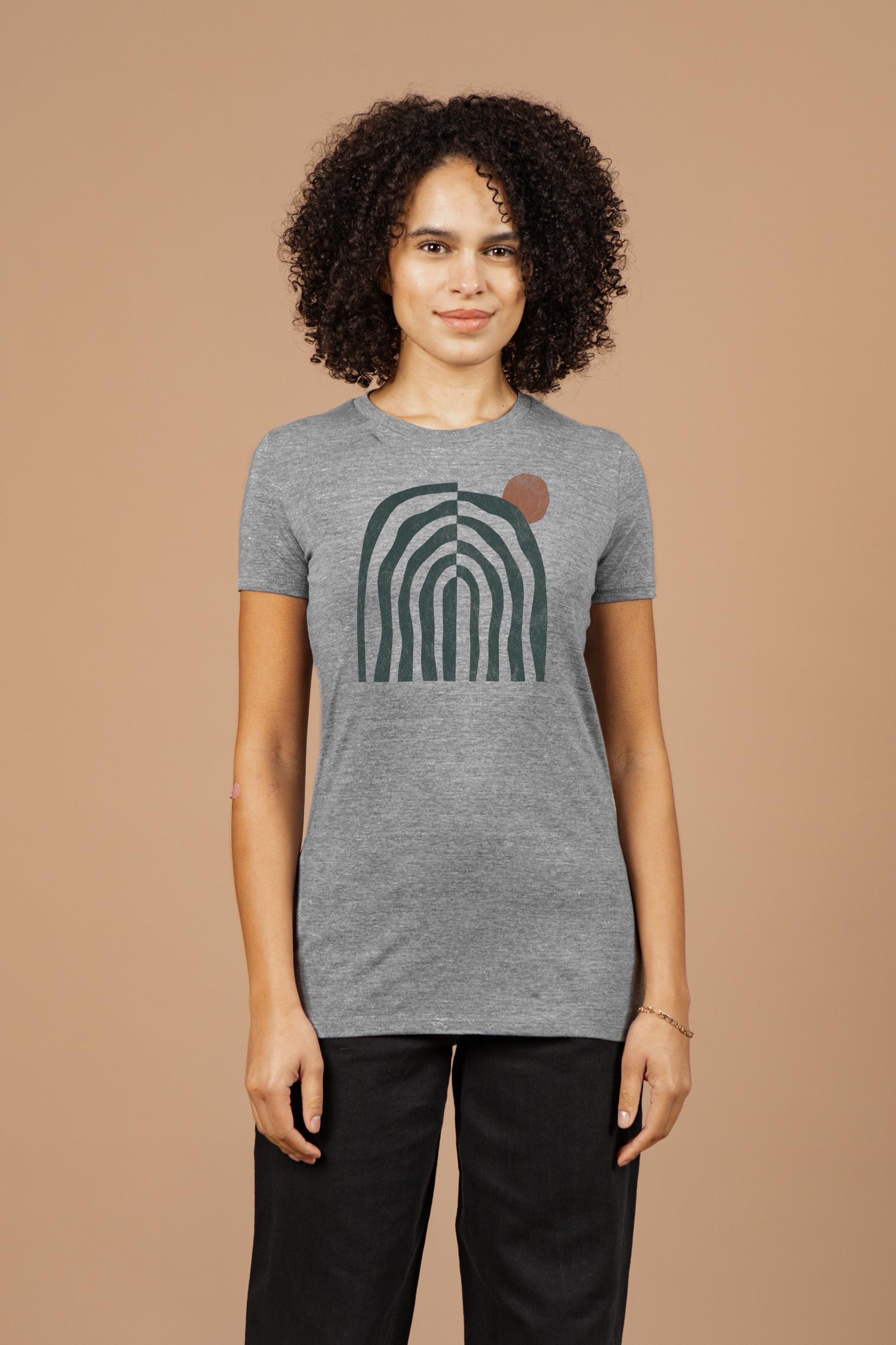 A person with curly hair wears the Bridge & Burn Women's Canopy Tee in grey, featuring an abstract green arch and small brown circle, standing against a plain beige background.