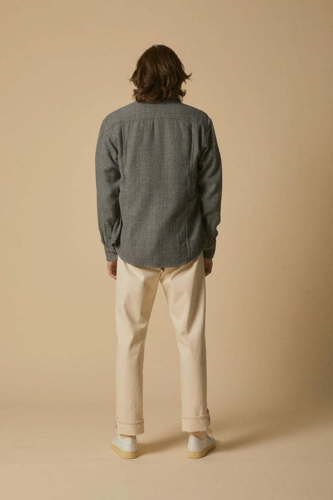 A person with medium-length brown hair, facing away, wears the Corridor Pearl Boucle Shirt in navy, cream pants, and light shoes against a plain beige background.