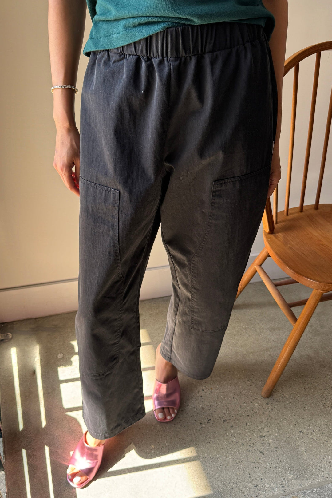 A person wearing Le Bon Shoppe's Gardener's Pant in Black, featuring a relaxed silhouette, and a green top stands next to a wooden chair. Sunlight casts shadows on the floor, accentuating the lightweight cotton-nylon fabric of the pants. Pink slide sandals complete the outfit.